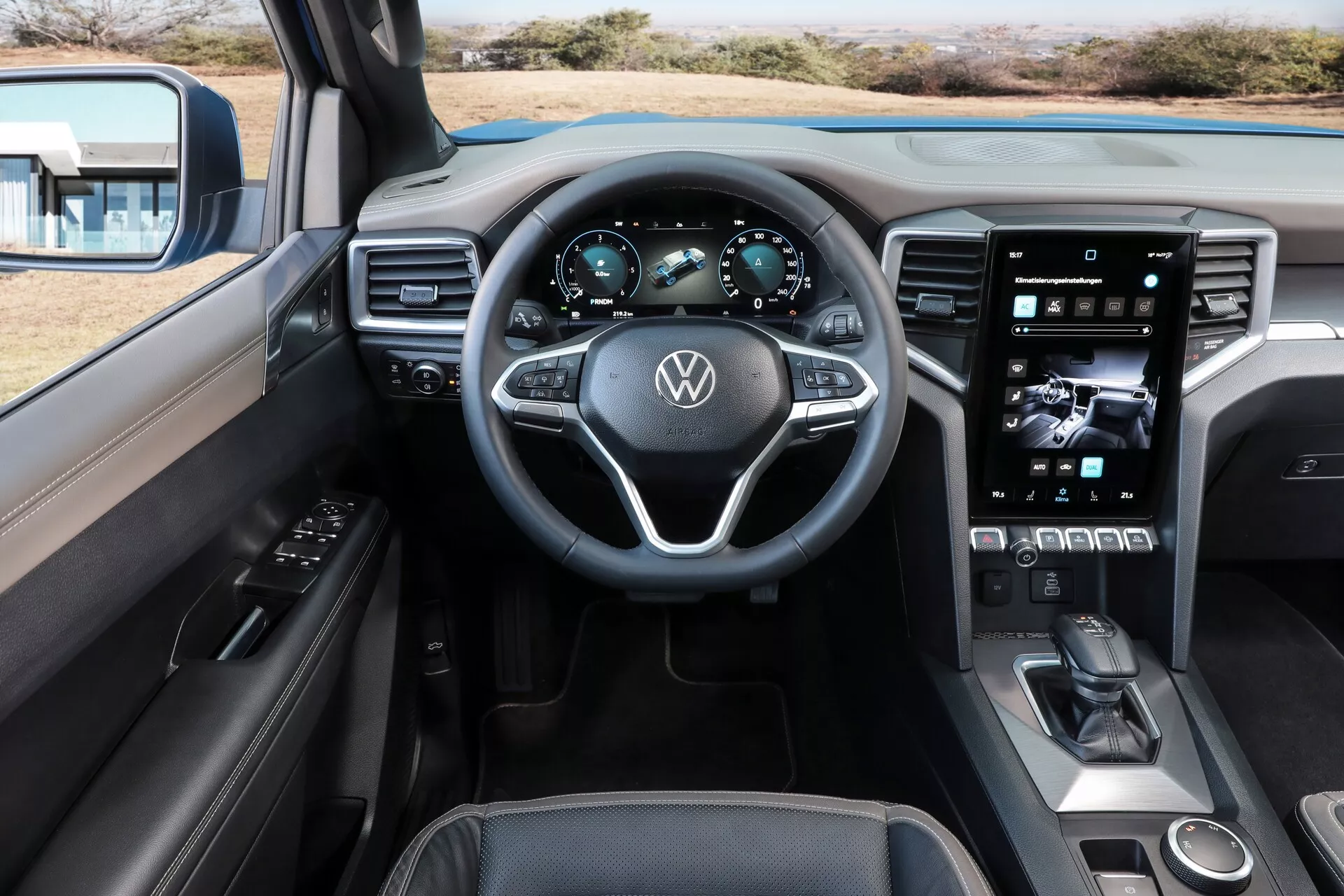 Notranji pogled kabine Volkswagen Amarok 2023 druge generacije iz zadnjega sedeža, ki prikazuje volan z logotipom Volkswagen, digitalno instrumentno ploščo, trikraki volan z integriranimi kontrolami, velik navpični zaslon na dotik za informacijsko-razvedrilni sistem, minimalne fizične kontrole, ki nakazujejo obliko, osredotočeno na zaslon na dotik, eleganten elektronski menjalni vzvod, izbirnik načina vožnje, možno območje brezžičnega polnjenja telefona, vrhunski materiali, vključno z usnjem in mehkimi na dotik površinami s podrobnimi šivi ter prilagodljivo ambientalno osvetlitvijo.