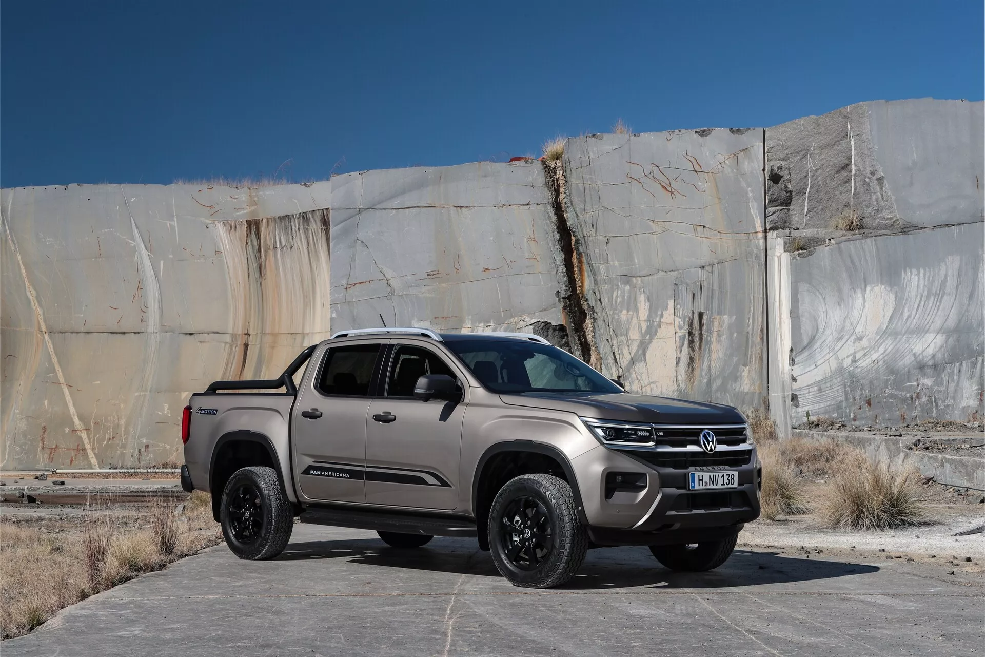 VW Nutzfahrzeuge Amarok 2023 druge generacije pickup v zunanjem okolju, rahlo postavljen v desni del okvirja z industrijsko ali zapuščeno stavbo v ozadju, ki ima velike razpokane betonske plošče. Posnetek iz tričetrtinskega sprednjega pogleda na nivoju oči poudarja sprednjo rešetko, žaromete verjetno z LED lučmi, pokrov motorja in stranski profil, vključno s sprednjim kolesom in vrati na strani voznika. Vozilo ima opazno sprednjo rešetko z velikim okroglim znakom, robustna črna lita platišča, kovinsko barvo, večjo oddaljenost od tal, kar nakazuje zmogljivosti 4x4, in funkcije, kot so parkirni senzorji, stranska ogledala z vgrajenimi smerniki ter anteno na strehi. Scena je videti kot posneta v jutranji ali predvečerni svetlobi, ki poudarja oblikovalske linije in sence.