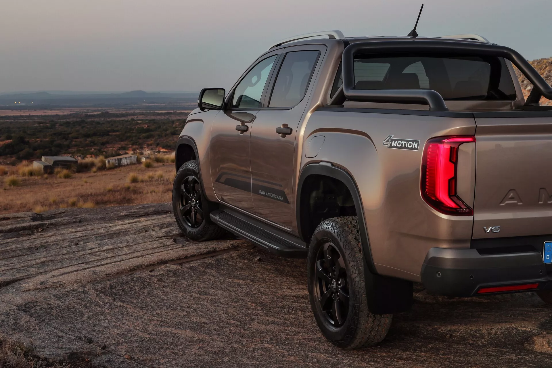 Slika VW Nutzfahrzeuge Amarok 2023 (2. generacije) prikazuje poltovornjak iz zadnje tričetrtinske perspektive, poudarja desno stran in zadnji del. Posnetek, narejen iz rahlo nagnjenega kota, poudarja tovorni prostor vozila, osvetljene zadnje luči, zadnji odbijač in vidne oznake znamke, modela ter morda nivo opreme ali specifiacijo motorja. Vozilo je postavljeno v grobo zunanjo okolje z vidno zemljo, ima kovinski premaz, športno palico v tovornem prostoru in črna kolesa, kar nakazuje izboljšano ali bolj športno opremo. Oznaka '4MOTION' kaže na sposobnost pogona na vsa kolesa ali štirikolesni pogon, medtem ko logotip na zadnjem pokrovu in vgrajena stopnica ali ročaj kažeta na tehnologijo enostavnega dostopa do tovornega prostora.