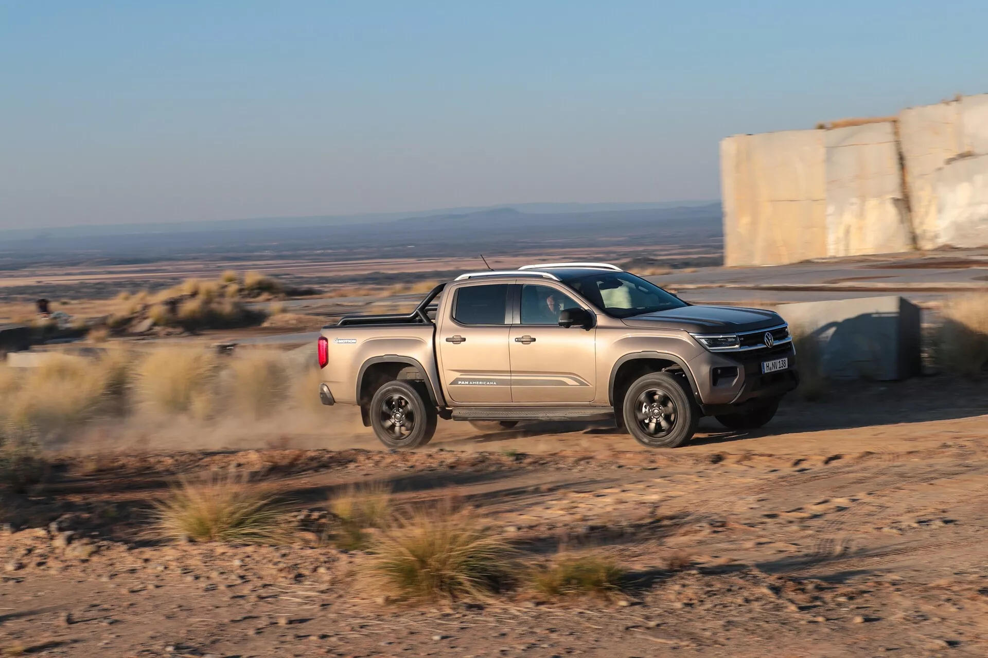 Poltovornjak Volkswagen Nutzfahrzeuge Amarok 2023 (2. generacija) ujet v gibanju na neasfaltirani poti, ki prikazuje svoje terenske sposobnosti s prašno sledjo za vozilom. Posneto pod poševnim kotom, poudarja stran za potnike, sprednji del in del kesona proti robustni pokrajini v poznih popoldanskih urah.