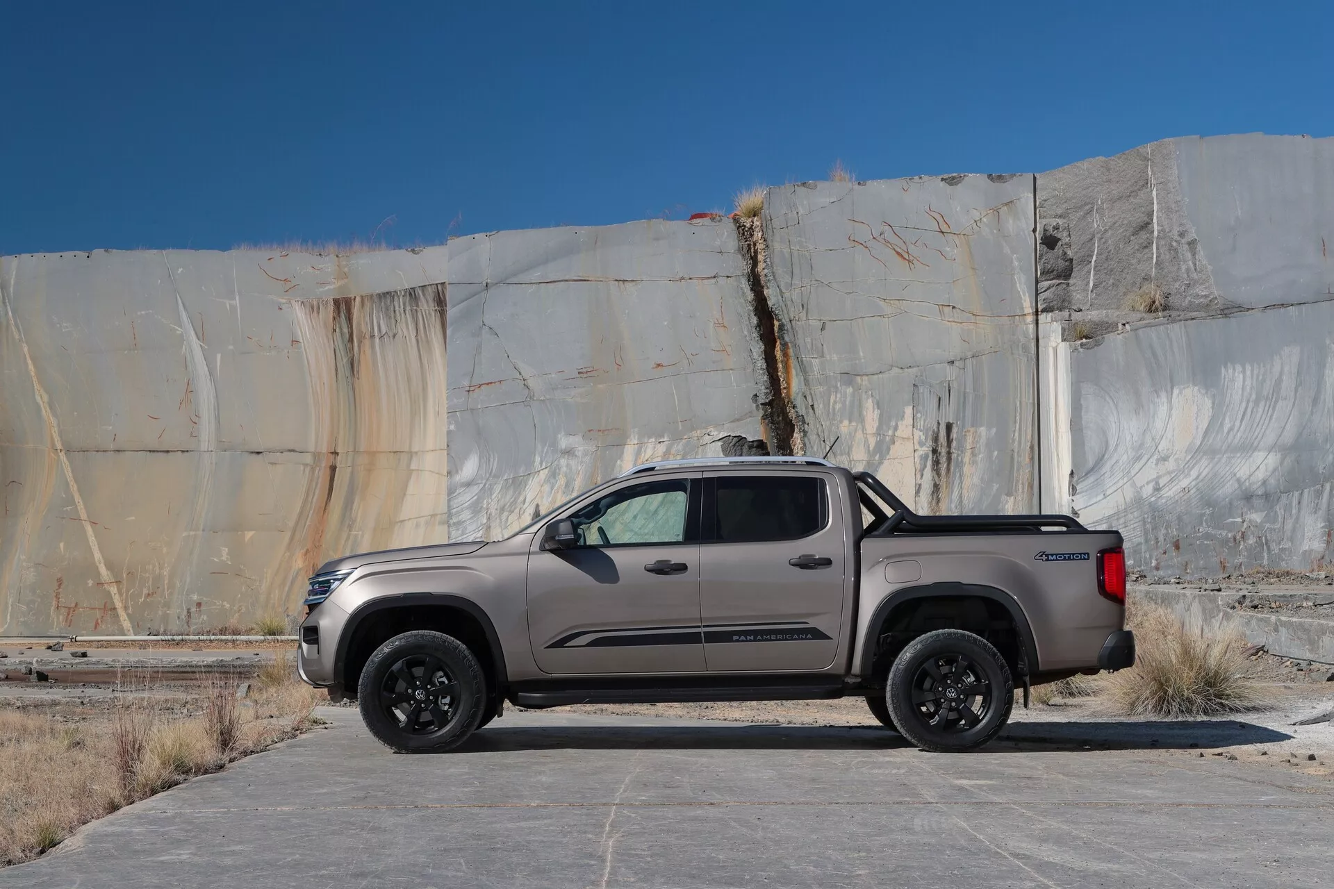 Slika prikazuje poltovornjak Volkswagen Amarok 2023 (2. generacija), parkiran na prostem, vzporedno s perspektivo kamere. Poltovornjak je sodobna dvokabinska kabina s štirimi vrati in kratko posteljo, gledano s strani, da je vidna celotna dolžina in višina. Vozilo je kovinske barve, bodisi sivo ali svetlo rjavo, z aerodinamičnimi linijami in elegantno karoserijo. Ima gladke krivulje, ki se zožijo proti zadnjemu delu, visoko pasovno linijo in zatemnjena zadnja okna. Poltovornjak stoji na črnih večkrakih kolesih, kar nakazuje višjo stopnjo opreme ali športni paket. Opremljen je s stranskimi stopnicami za lažji dostop ter z izrazitimi stranskimi ogledali, ki lahko vključujejo smerne kazalnike ali nadzor mrtvega kota. Ročaji vrat v barvi karoserije prispevajo k skladni estetski podobi. Predel s posteljo ima trdno oblogo za zaščito pred poškodbami tovora, zadnja vrata pa verjetno vključujejo tehnološke dodatke, kot je vzvratna kamera. Poltovornjak je parkiran na betonski ali asfaltni površini z lahkimi madeži pred strukturo z velikimi marmornimi ploščami, kar nakazuje industrijsko ali kamnolomsko ozadje. Dnevna svetloba z minimalnimi ostrimi sencami nakazuje oblačno nebo ali nadzorovano osvetlitveno okolje.
