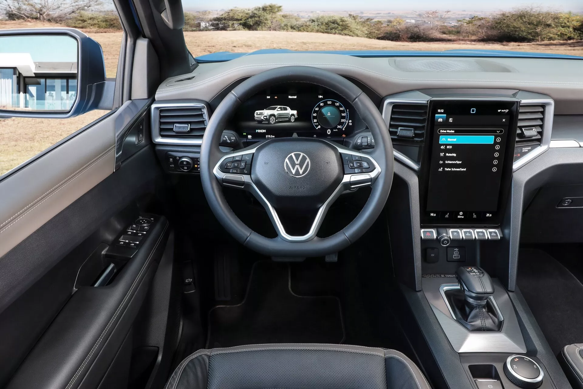 Notranji pogled Volkswagen Nutzfahrzeuge Amarok 2023 iz sredinskega zadnjega sedeža. Vidne značilnosti vključujejo tridimenzionalen večnamenski volan z logotipom VW in usnjeno prevleko, digitalno instrumentno ploščo, velik navpično usmerjen zaslon na dotik za informacijski sistem, minimalne fizične kontrole, elegantno klimatsko nadzorno ploščo in sodoben menjalnik z elektronsko parkirno zavoro. Delno je viden tudi voznikov vratni panel z nadzorom za okna in stranska ogledala. Notranjost kaže kombinacijo vrhunskih materialov, kot so usnje, mehke plastike in kovinski poudarki v temni dvobarvni kombinaciji. Sprednji sedeži so vidni z vgrajenimi vzglavniki in kontrastnimi šivi, kar nakazuje luksuzno končno obdelavo.