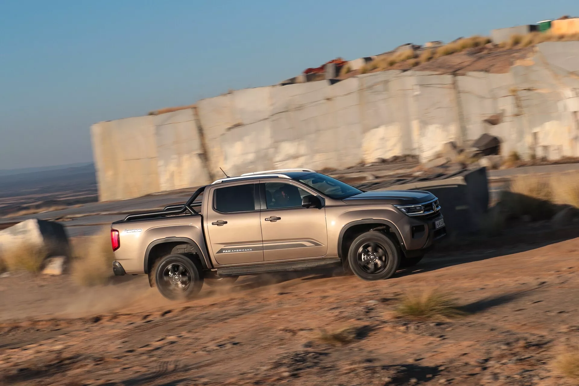 Stranski pogled fotografije pick-up tovornjaka VW Nutzfahrzeuge Amarok 2023 druge generacije v gibanju. Tovornjak se premika z leve proti desni v okviru v off-road okolju, prah se dviguje za zadnjimi kolesi. Topla, nizka sončna svetloba poudarja profil vozila, vključno z njegovimi robustnimi kolesi, modernimi LED žarometi in značilno sprednjo masko.