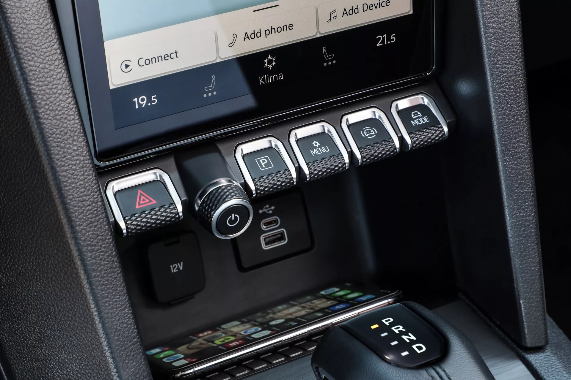 Posnetek osrednjega konzolnega območja v VW Nutzfahrzeuge Amarok 2023 druge generacije, ki prikazuje dvopodročno klimatsko napravo z zaslonom na dotik, prikazuje temperature 19,5°C in 21,5°C. Nad klimatskim nadzorom je viden rob informacijsko-zabavnega sistema, ki ponuja možnosti povezljivosti, kot sta 'Connect' in 'Add Phone.' Spodaj je pet kovinskih gumbov za opozorilne luči, parkirni senzorji, zagon/ustavitev motorja, načini vožnje ali nadzor oprijema ter nadzor spusta, poleg je 12V vtičnica. Na dnu je majhen prostor za shranjevanje ključev in denarnic. Na desni strani okvira je razviden samodejni menjalnik, ki prikazuje 'P' za parkiranje. Materiali in obdelave odražajo premium ter moden dizajn.