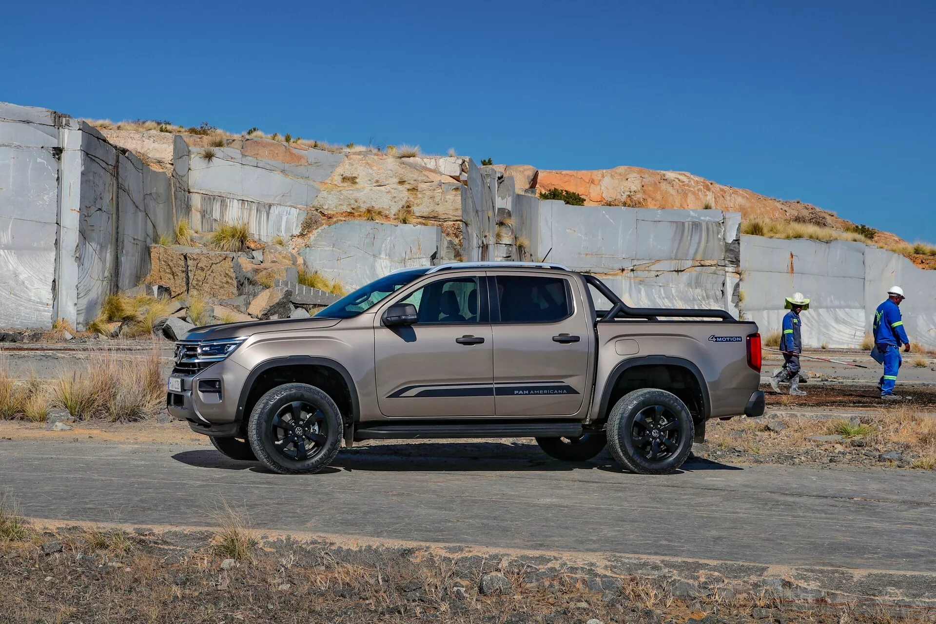 Slika prikazuje štirivratni pickup VW Nutzfahrzeuge Amarok 2023 druge generacije, ki se nahaja na prostem s stranskim profilom proti kameri. Vozilo je fotografirano iz rahlo nizkega kota, kar poudarja njegovo oddaljenost od tal in robustno gradnjo, idealno za različne terene. Tovornjak je parkiran vzporedno s perspektivo kamere, kar omogoča jasen pogled na njegovo stran, ki vključuje sprednja in zadnja vrata potnikov, stranska ogledala, ročaje, oblikovne linije, koloteke in kolesa. Vozilo se nahaja na ravnem, neasfaltiranem površju, ki spominja na gradbišče, zaprašen teren in delavci v ozadju pa potrjujejo njegovo uporabnost za industrijsko ali komercialno rabo. Ključna področja v središču pozornosti na sliki so sposobnost terenske vožnje, oblikovanje vzmetenja, paket koles in pnevmatik ter zaključna obdelava barve in stopnja opreme vozila.