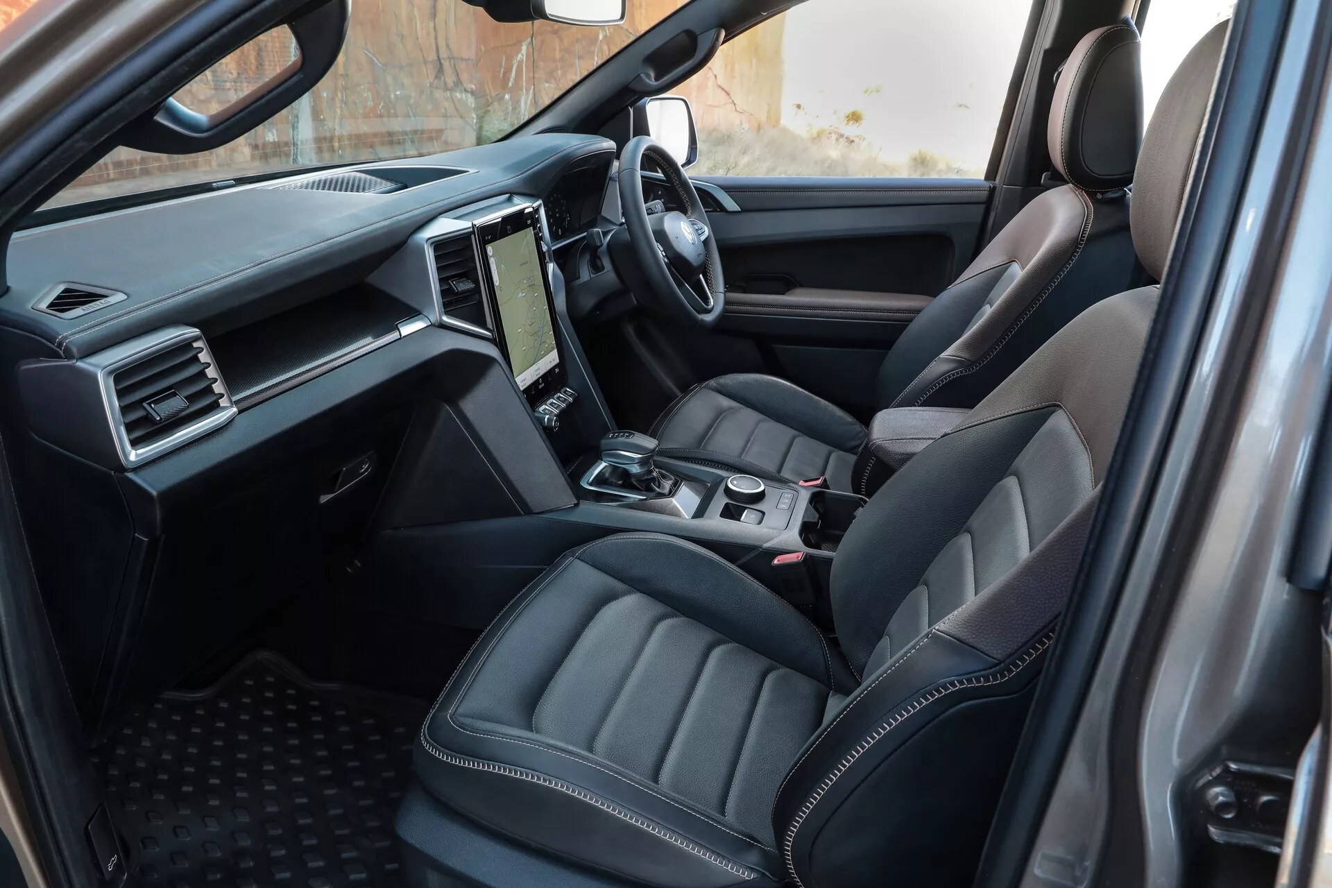 Notranji pogled VW Nutzfahrzeuge Amarok 2023 (2. generacija), posnet iz perspektive zadnjega potnika, z poudarkom na voznikovem konturiranem usnjenem sedežu, osrednji konzoli z nadzornimi elementi in moderni armaturni plošči z zaslonom na dotik za infotainment sistem.