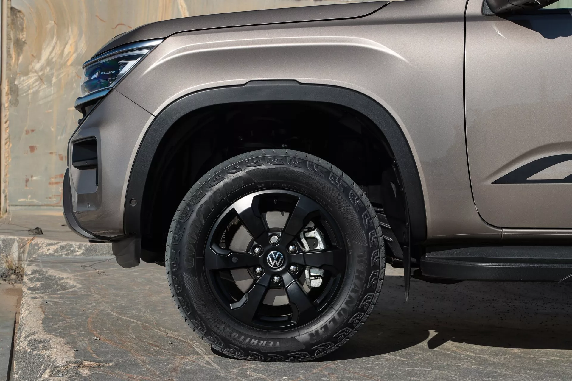 Pogled od blizu na sprednje kolo Volkswagen Amarok 2023 (2. generacija), z večkrakom sijoče črnim kolesom in terensko pnevmatiko, mat/satenastim zaključkom barve, plastičnim blatniškim podaljškom, bočno stopnico pod potniškimi vrati ter delno vidno LED žarometom in oblikovano karoserijsko linijo.