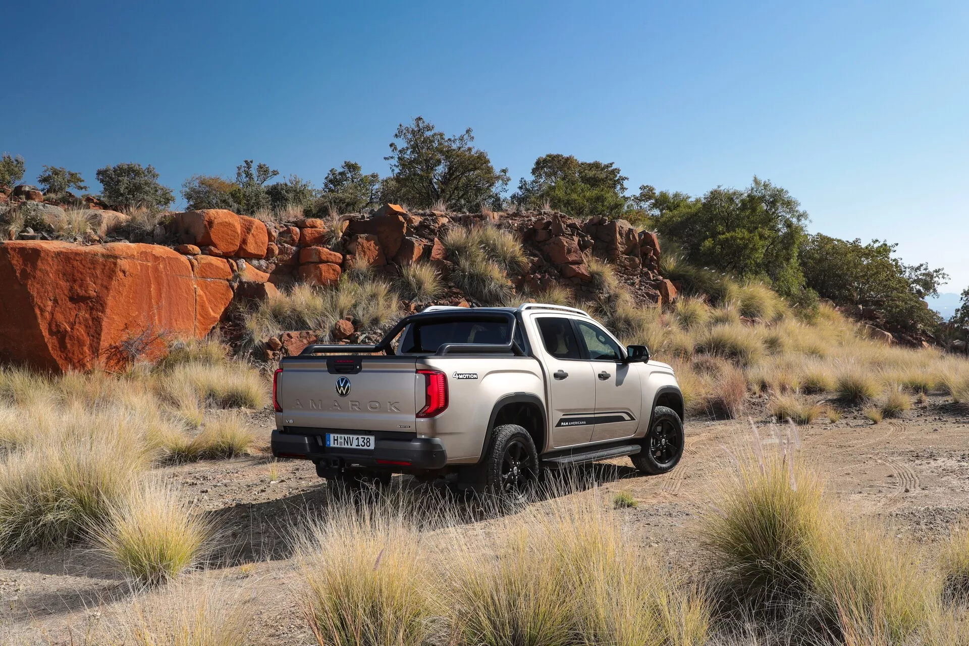 Slika pick-up vozila Volkswagen Nutzfahrzeuge Amarok 2023, parkiranega na prostem na makadamski cesti z naravno skalnato pokrajino v ozadju, posneta iz polovične zadnje perspektive. Elegantno vozilo s kromiranimi poudarki, osvetljeno z naravno svetlobo, prikazuje športno in robustno zasnovo z dvojno kabino, kratko posteljo, izrazitimi blatniki in debelimi pnevmatikami, primernimi za terenske avanture.
