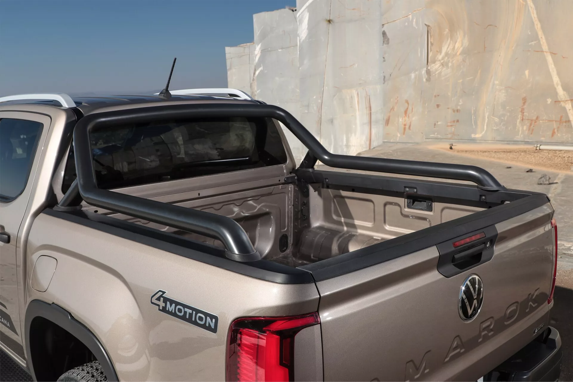 Bližji, kotni pogled na zadnji del bež ali svetlo zlatega tovornjaka Volkswagen Amarok 2023 (2. generacija). Posnetek se osredotoča na tovorni prostor in zadnja vrata, kjer so prikazani logotip Volkswagen in ime Amarok, vtisnjeno v kovino. Vidne lastnosti vključujejo kratki tovorni prostor s plastično oblogo, zadnjo svetlobno enoto na desni strani, značko 4MOTION za štirikolesni pogon in stopnico na odbijaču za lažji dostop. Zadnje okno, verjetno z odmrzovalnikom, omogoča vpogled v čisto in dobro vzdrževano kabino za potnike. Slika zajema oblikovane linije karoserije, ki kažejo na izpopolnjeno avtomobilsko zasnovo ob dnevni naravni svetlobi.