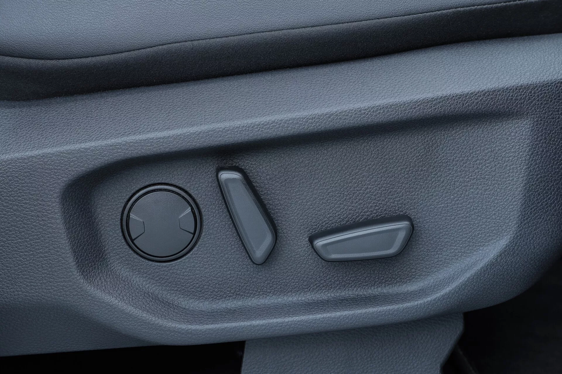 Bližnji posnetek notranje vratne plošče VW Nutzfahrzeuge Amarok 2023, s tremi zavitimi stikali za nastavitev vzvratnih ogledal, dvema navpično poravnana stikaloma verjetno za električna okna in nadzor zaklepanja vrat ter teksturirano površino temnega odtenka, ki nakazuje trajen, nedrseč material. Fotografija je posneta pod rahlim kotom navzdol, da jasno prikaže kontrole.
