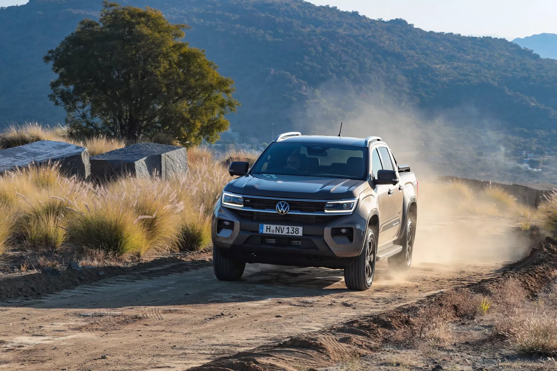 Profesionalna fotografija druge generacije poltovornjaka Volkswagen Nutzfahrzeuge Amarok 2023, posneta s treh četrtin spredaj. Slikaje izpostavlja moderno in robustno sprednjo stran vozila z izrazito masko in logotipom Volkswagen ter LED dnevnimi lučmi. Amarok je v gibanju po prašni ali gramozni cesti v naravnem okolju, kjer zadnja kolesa dvigujejo prah, kar kaže na terensko uporabnost in zmogljivost na vseh terenih. Aluminijasta kolesa, velika oddaljenost od tal in vzmetenje prispevajo k zmogljivosti in trajnosti vozila, kar je pomembno za kategorijo SUV/off-road. Nizek kot posnetka poudarja dominanten in močan videz vozila.