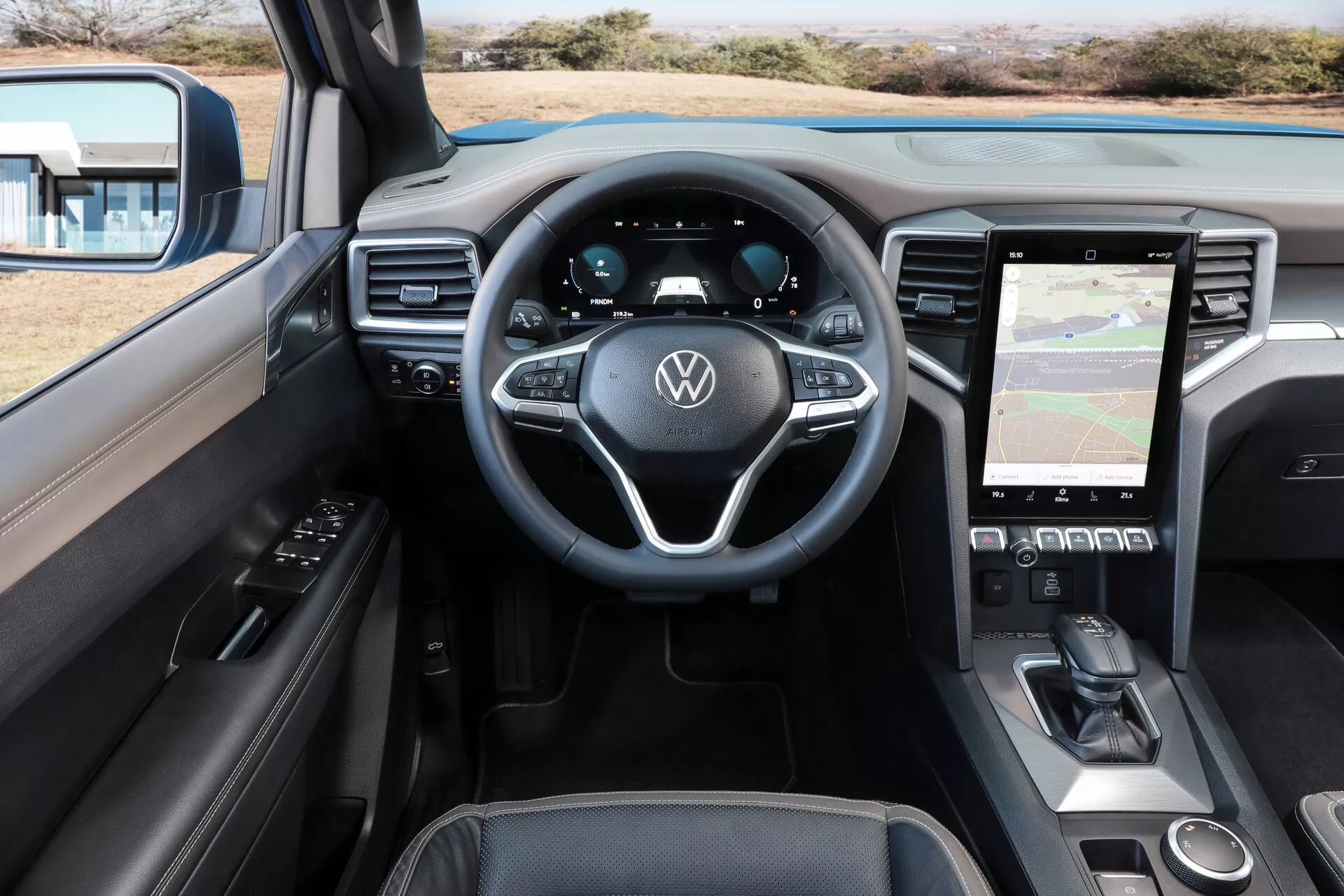Pogled na notranjost kabine VW Nutzfahrzeuge Amarok 2023 (2. generacija) iz perspektive voznika. Slika je posneta iz nekoliko dvignjenega kota, ki prikazuje sprednji del kabine. Viden je volan z logotipom Volkswagen in večfunkcijskimi kontrolami, popolnoma digitalni instrumentni zaslon in navpično usmerjen infotelematični sistem na dotik, ki prikazuje zemljevid. Sredinska konzola vključuje nadzor klimatske naprave, morebitno območje za brezžično polnjenje in avtomatsko prestavno ročico. Elegantna in sodobna zasnova vključuje usnjene in mehke materiale na armaturni plošči in vratnih panelih, z gumbi za upravljanje oken in nastavitvami ogledal na voznikovih vratih.