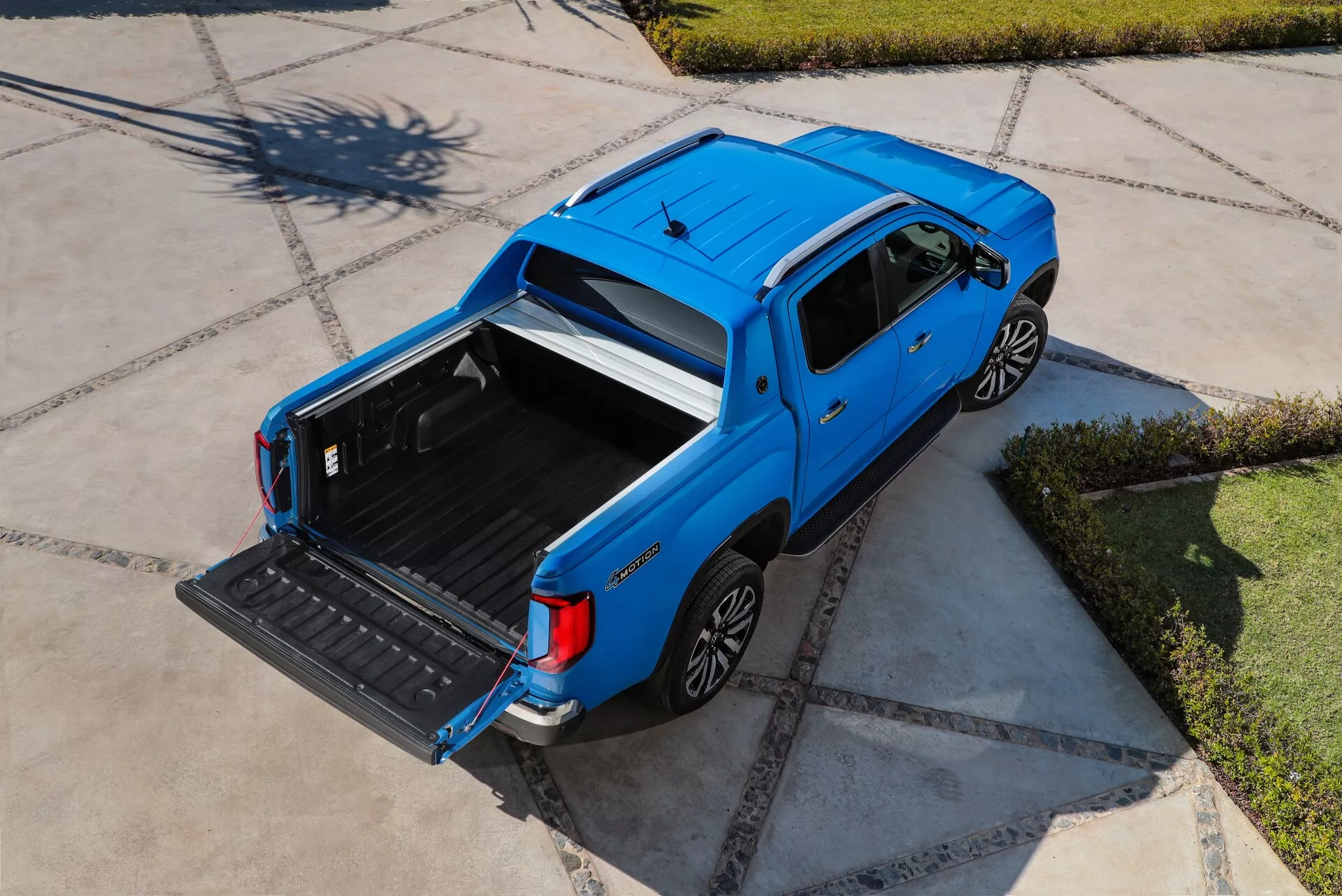 Pogled iz zraka na modri pick-up VW Nutzfahrzeuge Amarok druge generacije z letom 2023. Slika prikazuje vozilo s posadkovsko kabino, polno velikimi zadnjimi sedeži in štirimi vrati. Tovorni prostor je odprt in prazen, z odprtim prtljažnikom, kar poudarja uporabnost vozila. Vidne so elegantne linije karoserije, aluminijasta kolesa v vidnih lokih koles in kljuke v barvi karoserije. Okolje se zdi stanovanjsko, z dobro vzdrževanim pločnikom, pod jasnim nebom z visokim soncem.