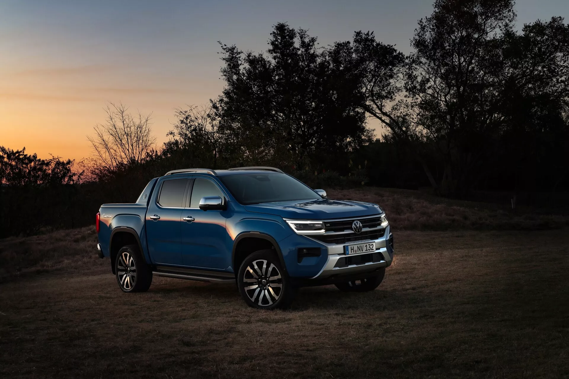 Stranski profil modrega VW Nutzfahrzeuge Amarok 2023 (2. generacija) pick-upa med mrakom ali zoro, ki prikazuje stran vozila, sprednjo rešetko in žaromete z oranžnim obzorjem in slabo osvetljenim okoljem. Oblikovne značilnosti vozila, vključno s sodobnimi LED žarometi, kromirano sprednjo rešetko, vgrajenimi smerniki v stranskih ogledalih, oblikovanjem aluminijastih platišč in sijočim zaključkom barve, so poudarjene s poudarjeno umetno osvetlitvijo. Prizor uravnoteži pustolovski, robusten značaj pick-upa z naravnim zunanjim okoljem in poudarja njegovo vsestranskost in zmogljivost v terenskih pogojih.