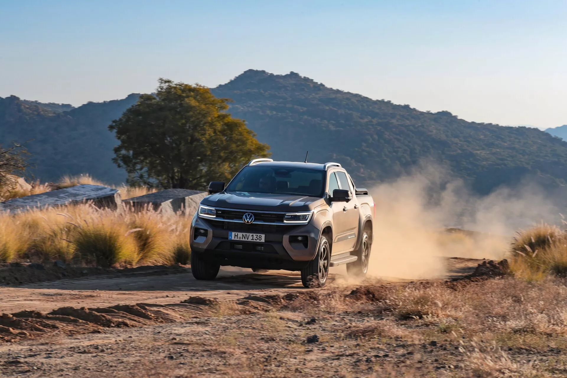 Slika prikazuje VW Nutzfahrzeuge Amarok 2023 druge generacije, ki vozi po neasfaltirani poti, verjetno makadamski ali gramozni poti, v podeželski ali gorski pokrajini. Vozilo je fotografirano s tridčetrtinskega sprednjega kota, kar prikazuje njegovo rešetko, odbijač, žaromete, meglenke, pokrov motorja, sprednje blatnike, kolesa in stranski profil. Mehka osvetlitev, verjetno zjutraj ali pozno popoldne, poudari oblak prahu, ki ga povzroči vozeče vozilo, kar kaže na njegovo hitrost in suhe razmere na terenu. Oblikovanje vozila, kot so kolesa, primerna za terenske razmere, robusten sprednji odbijač in napredna razsvetljava, nakazujejo sodobne funkcije in tehnologijo, vključno s kontrolo oprijema, stabilnostjo v zahtevnih pogojih in morebitno naprednimi sistemi za pomoč vozniku.