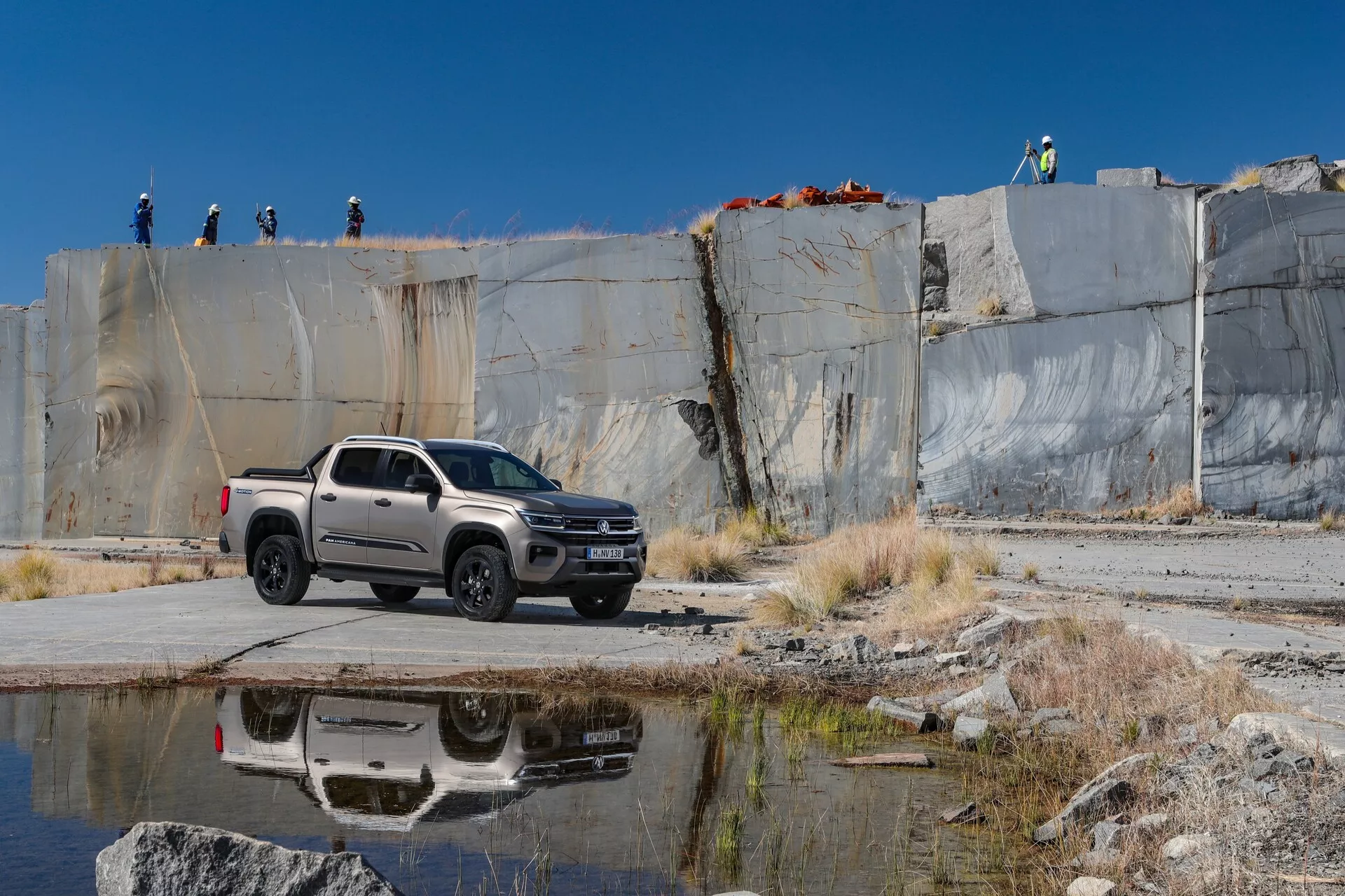 Slika prikazuje pickup vozilo VW Nutzfahrzeuge Amarok 2023 druge generacije, parkirano na betonski ali asfaltirani površini v kamnolomu ali gradbišču, ujeta iz treh četrtin sprednjega kota. Prednja stran vozila, vključno s žarometi, rešetko in odbijačem, ter bočni profil s kolesi, vrati in stranskimi ogledali, so jasno vidni. Vozilo ima sodoben dizajn z robustnimi linijami karoserije, konfiguracijo kabine za posadko in krajšo tovorno površino, barvano v svetlo barvo, verjetno srebrno ali sivo. Naravna sončna svetloba poudari oblikovne značilnosti in barvo. V ozadju so vidni ljudje s čeladami na dvignjenem robu, kar kaže na aktivno industrijsko ali gradbeno delovno mesto. Vidne tehnološke lastnosti vključujejo aluminijasta platišča, stranska ogledala z vgrajenimi smerniki in LED žaromete. Odsev vozila na bližnji vodni površini dodaja estetsko dimenzijo in nakazuje njegove terenske sposobnosti.
