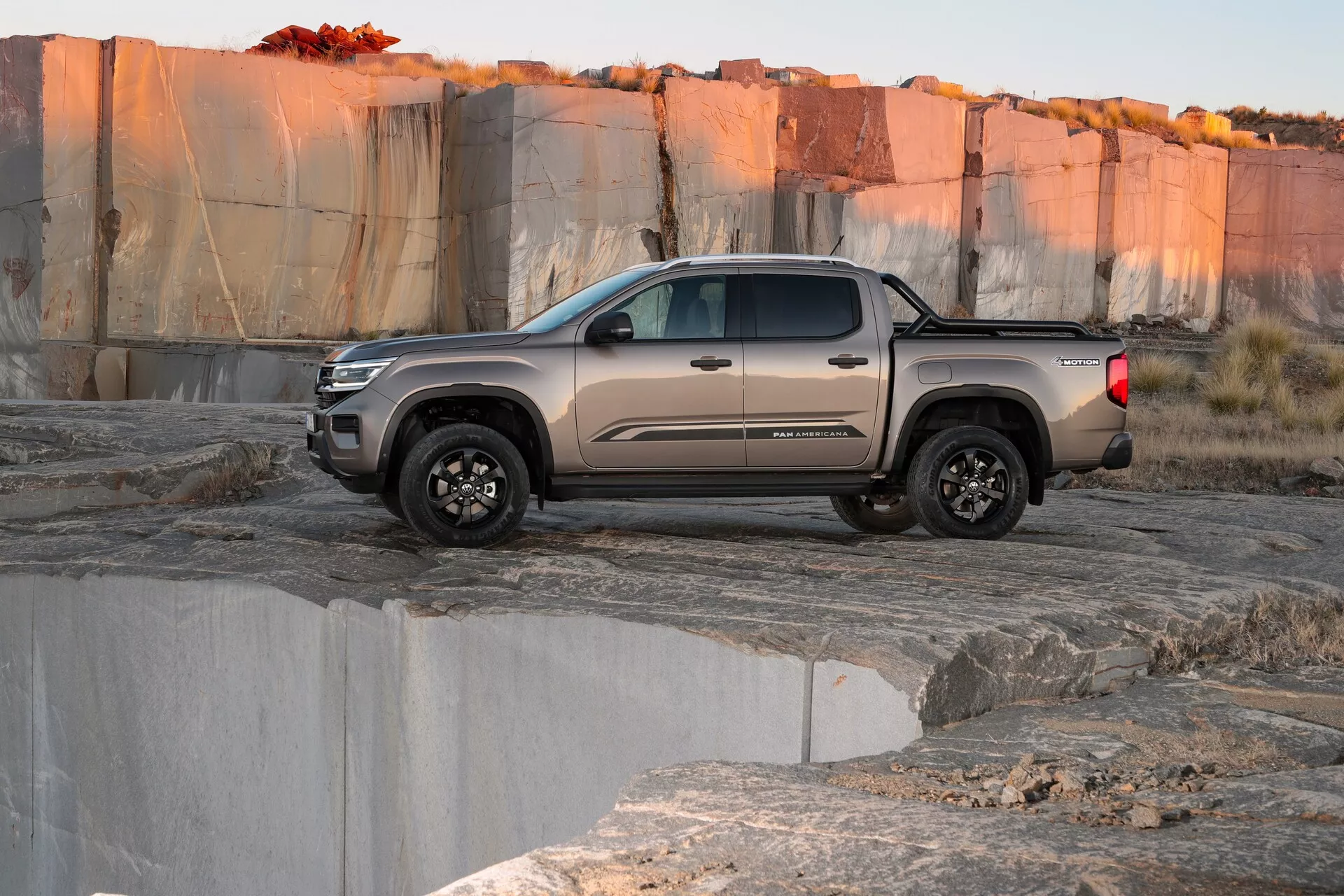 Stranski pogled na drugo generacijo VW Nutzfahrzeuge Amarok 2023, srednje velik poltovornjak s konfiguracijo kabine s posadko in terenskimi pnevmatikami, postavljen na kamnit, neraven teren, ki spominja na kamnolom ali naravno okolje ob sončnem vzhodu ali zahodu. Nizkokotni posnetek poudarja višino vozila od tal in robustno zmožnost. Vozilo je v zadržanem taupe ali sivem tonu s temnimi kolesi, zajemajoč bistvo sodobnega avtomobilskega oblikovanja.