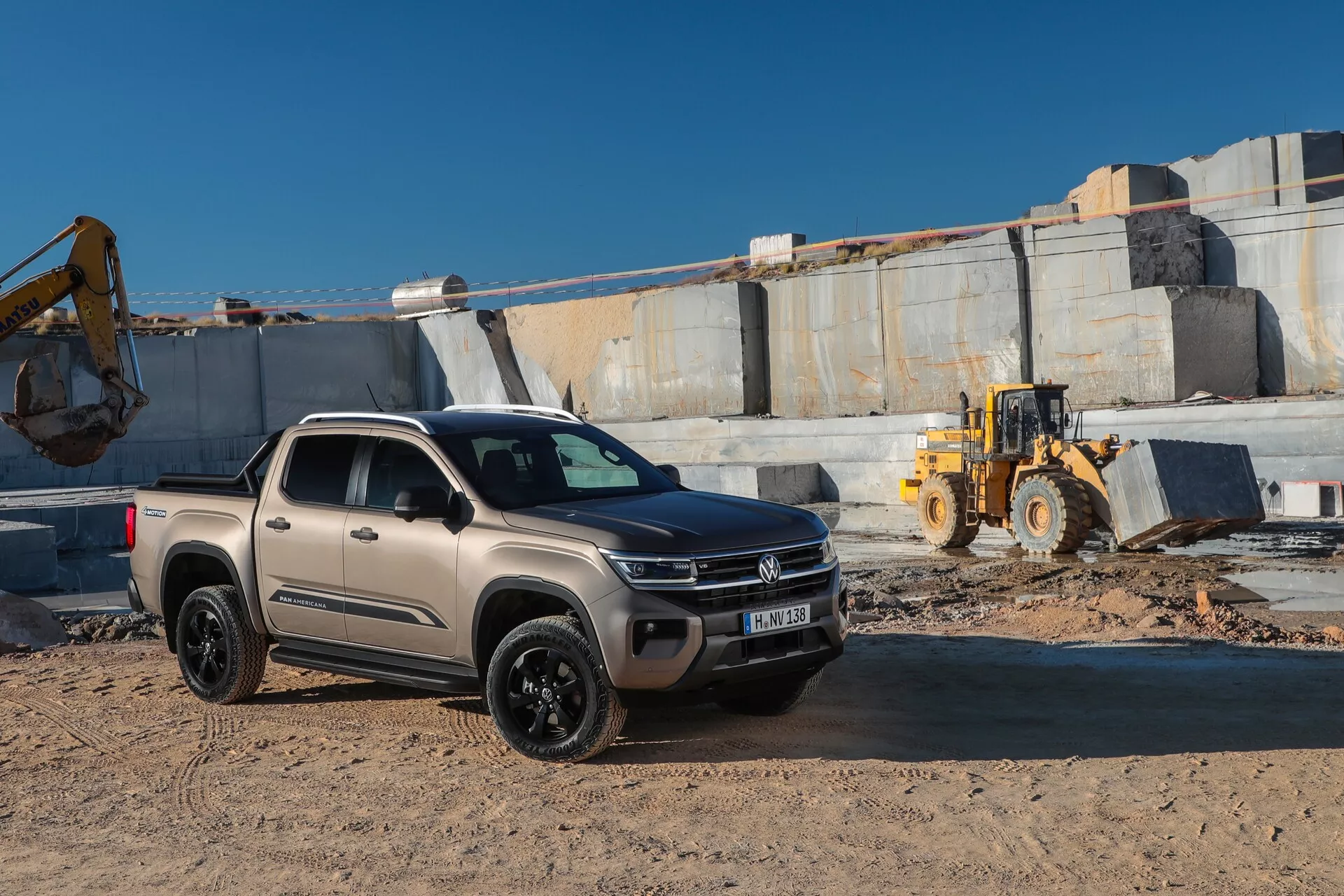 Poltovornjak VW Nutzfahrzeuge Amarok 2023 druge generacije je parkiran na gradbišču z elementi kamnoloma v ozadju. Fotografija je posneta iz nizkega kota, prikazuje prednjo masko, žaromete, pokrov motorja in stranski profil na sovoznikovi strani. Ima dvojno kabino, moderne oblike z gladkimi linijami, robusten videz s terenskimi pnevmatikami in povečano oddaljenostjo od tal. Barva tovornjaka je mat, verjetno bež ali svetlo taupe barve, s črnimi kolesi in LED žarometi z dnevnimi lučmi. Okolje vključuje težko mehanizacijo in prašno, neravno površino.