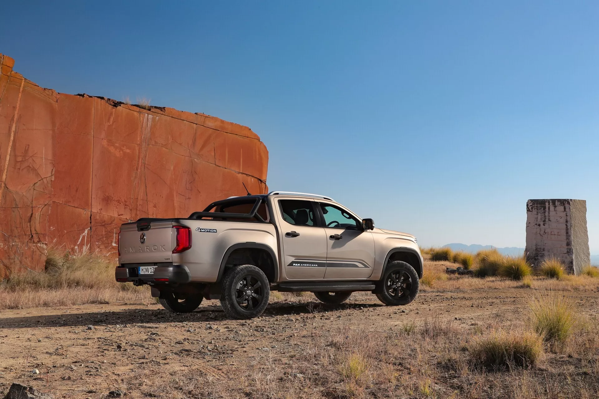 Bočni pogled na pickup VW Nutzfahrzeuge Amarok 2023, posnet iz rahlo zadnjega kota, parkiran na suhem, ravnem puščavskem terenu z redko vegetacijo in jasnim modrim nebom, proti ozadju velikih rdečih skalnatih formacij. Stranski profil tovornjaka, vključno s sprednjo mrežo, žarometi, vozniškimi vrati, sopotnikovim profilom, tovornim prostorom in zadnjimi vrati, so popolnoma vidni. Značilne lastnosti, kot so stranska ogledala, dizajn koles in stranska stopnica, prispevajo k robustnemu videzu. Nizki kot posnetka poudarja oddaljenost od tal in močan položaj, s sencami, ki nakazujejo pozno popoldansko osvetlitev.