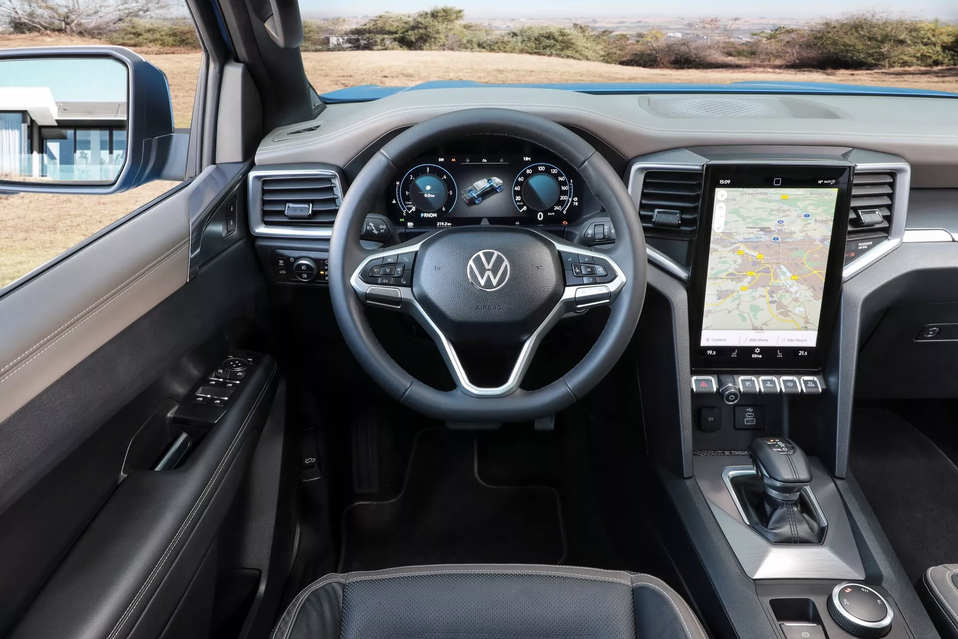 Notranji pogled Volkswagnovega vozila Nutzfahrzeuge Amarok 2023, ki prikazuje perspektivo voznika z vidika armaturne plošče, volana in osrednje konzole. Volan ima trikraki dizajn z večfunkcijskimi kontrolami in logotipom Volkswagen. Digitalna instrumentna plošča voznika prikazuje merilnik hitrosti, tahometer, merilnik goriva in morda navigacijske ali potovalne informacije. Osrednja konzola vsebuje velik zaslon na dotik, ki prikazuje navigacijski zemljevid, spodaj pa so nadzorne plošče za klimatske naprave na dotik. Notranjost nakazuje napredno tehnologijo v vozilu in sodoben dizajn z mehkimi površinami, kovinskimi poudarki in usnjenim oblazinjenjem.