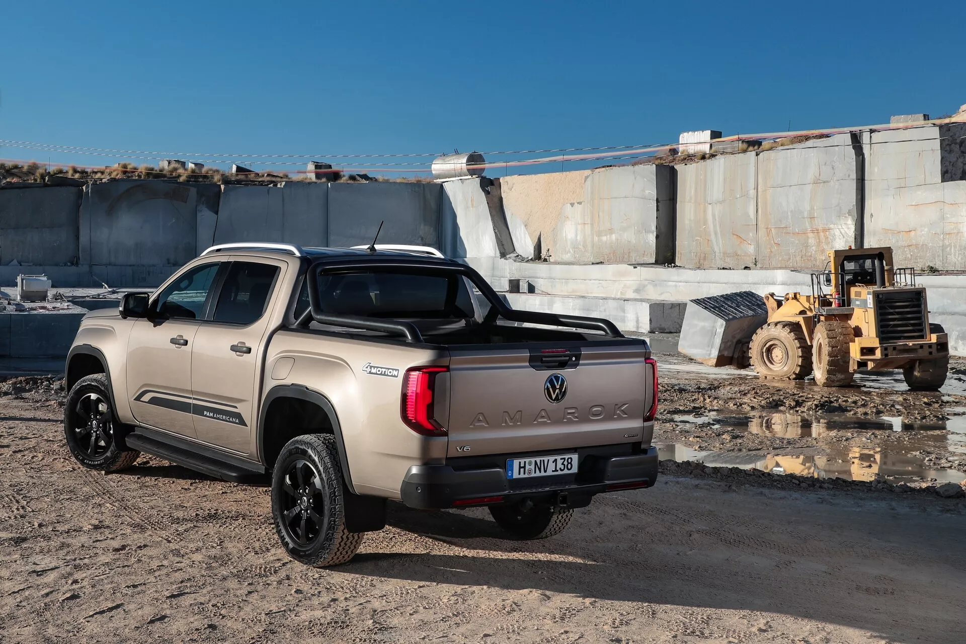 Ta slika prikazuje Volkswagen Amarok 2023 (druga generacija) pick-up iz perspektive zadnje tri-četrtine, s poudarkom na desni strani vozila. Tovornjak je parkiran na gramozni površini v osvetljenem zunanjskem okolju, verjetno na gradbišču ali v industrijskem območju, kar nakazuje prisotnost težke mehanizacije v ozadju. Ključne značilnosti vključujejo drzen napis 'AMAROK' na zadnjih vratih, športno palico na postelji, horizontalne zadnje luči ter temna aluminijasta platišča z več kraki. Svetlo bež barva in konfiguracija kabine z dvema deloma s štirimi polnovrednimi vrati nakazujeta prostorno kabino. Registrska tablica v spodnjem levem kotu je zamegljena zaradi zasebnosti.