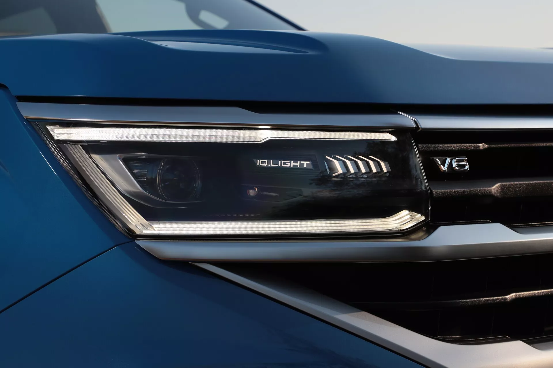 Bližnji posnetek sprednjega levega žarometa modernega VW Nutzfahrzeuge Amarok 2023 (2. generacija). Posnetek je narejen iz nizkega kota, poudarjajoč elegantno, napredno IQ.LIGHT LED tehnologijo z značilnim cikcak vzorcem. Emblem V8 na mreži je delno viden, kar nakazuje visokozmogljiv motor. Barva avtomobila je temnejšega odtenka, verjetno modra, v naravni svetlobi, poudarjajoč konture in tridimenzionalno oblikovanje vozila.