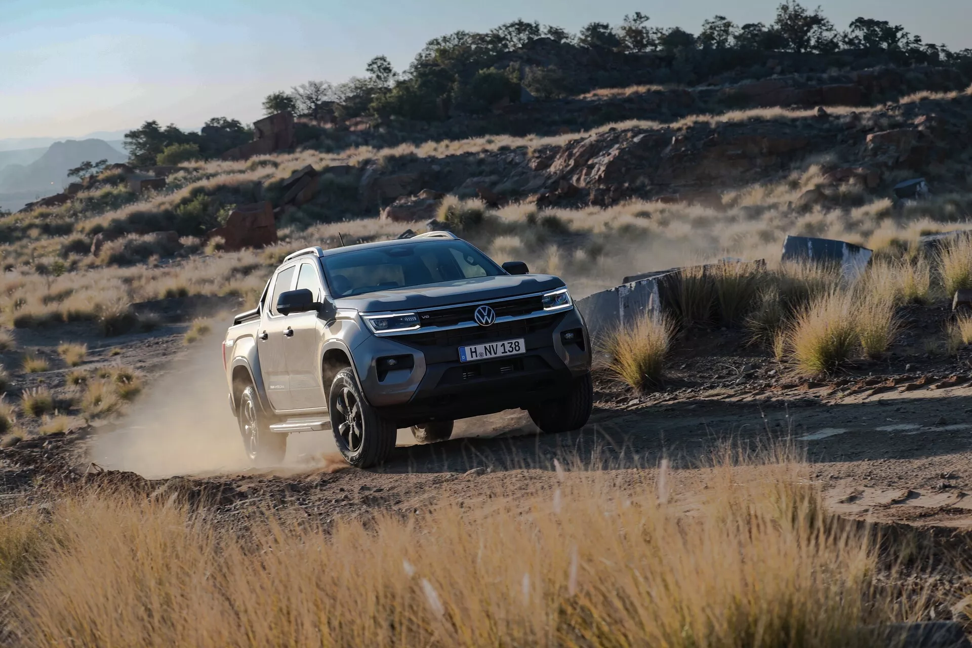 Dinamičen posnetek druge generacije SUV VW Nutzfahrzeuge Amarok 2023 v terenskem okolju. Sliko zajema trikratni sprednji bočni pogled, ki izpostavlja rešetko vozila, žaromete, sprednji odbijač in pokrov motorja ter voznikovo stran od sprednjega blatnika do zadnjih vrat. SUV, pobarvan v svetlo barvo, kontrastira s grobim ozadjem prašnih poti, suhe trave in skalnatega terena. Povišana oddaljenost od tal in velika lita platišča poudarjajo njegove terenske zmogljivosti. Oblak prahu, ki ga dvigujejo zadnje pnevmatike, nakazuje hitro gibanje. LED žaromete in dnevne luči so jasno vidni, kar poudarja sodobno svetlobno tehnologijo. Znamenje proizvajalca, oblika rešetke in tekstura prahu so vidni, kar poudarja eleganten dizajn in robusten videz vozila.