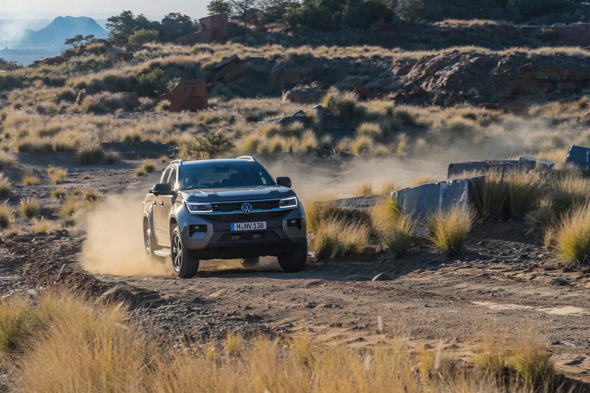 Volkswagen Amarok SUV 2023 med vožnjo po makadamski cesti, tričetrtinski pogled s sprednje strani prikazuje LED žaromete, podroben sprednji del z emblemom in obliko koles na grobem terenu z oblakom prahu.