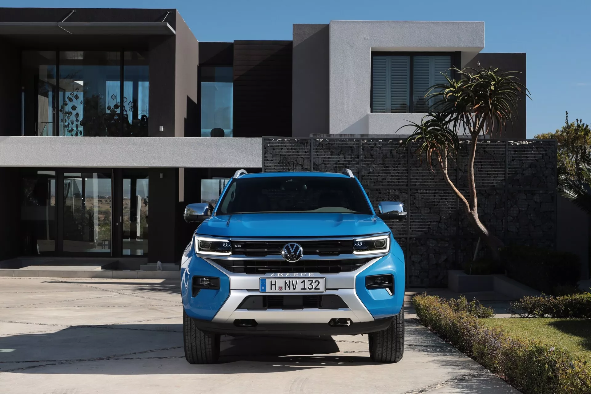 Sprednji pogled modrega Volkswagna 2023 Amarok SUV, parkiranega pred sodobno stanovanjsko zgradbo, poudarja elegantne žaromete vozila, dizajn mreže, dnevne luči, izpostavljen logotip Volkswagen, senzorje na spodnjem odbijaču in radar, kar poudarja zasnovo, razsvetljavo in varnostne tehnologije.