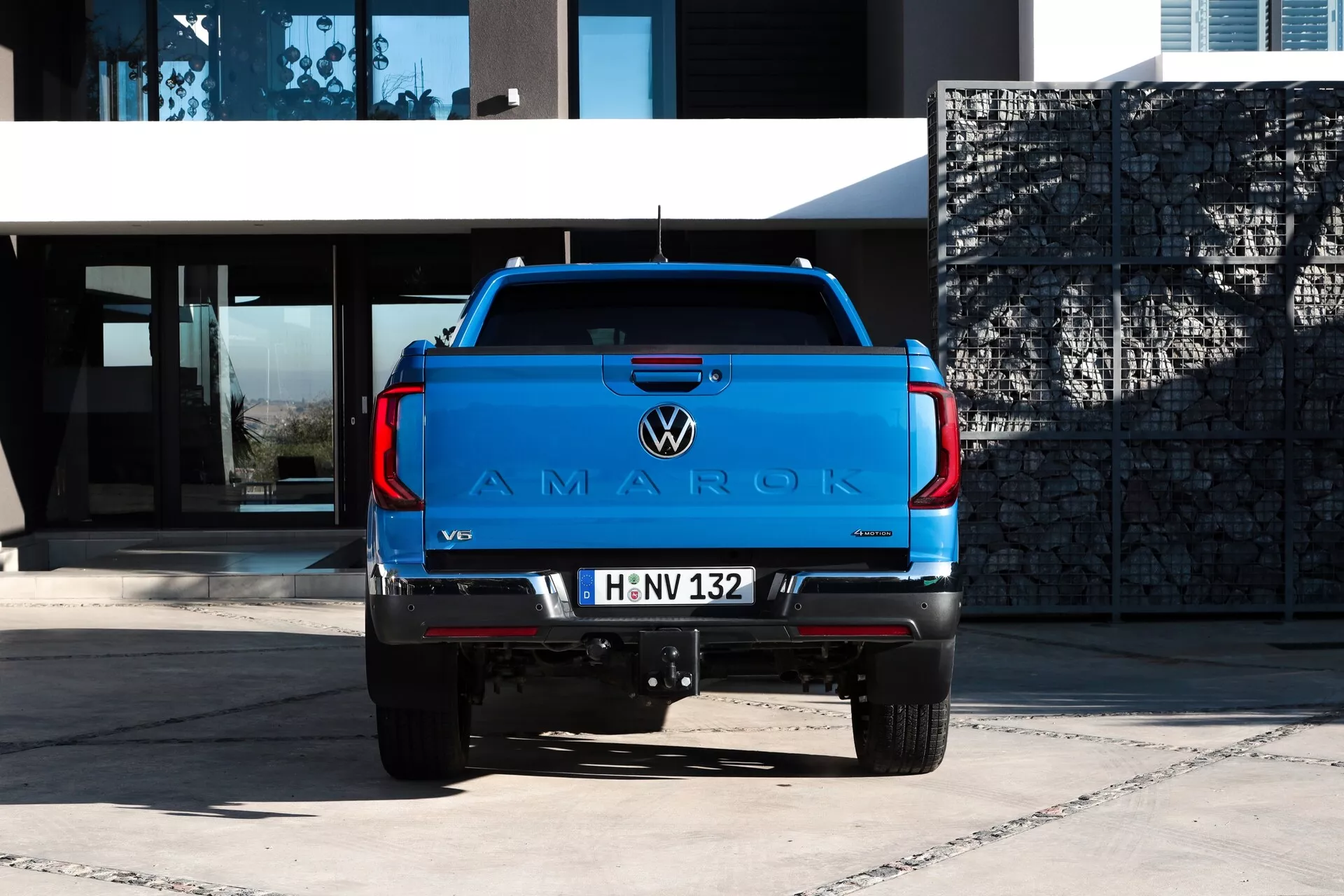 Zadnji pogled na Volkswagen Amarok 2023 pickup s sijočo modro barvo na asfaltu, ki prikazuje velik logo 'AMAROK' na zadnjih vratih, VW emblem, značko motorja V6, sodobne zadnje luči, vlečno kljuko, parkirne senzorje, evropsko tablico in dvojni izpušni sistem, v ozadju moderne stavbe v naravni dnevni svetlobi.