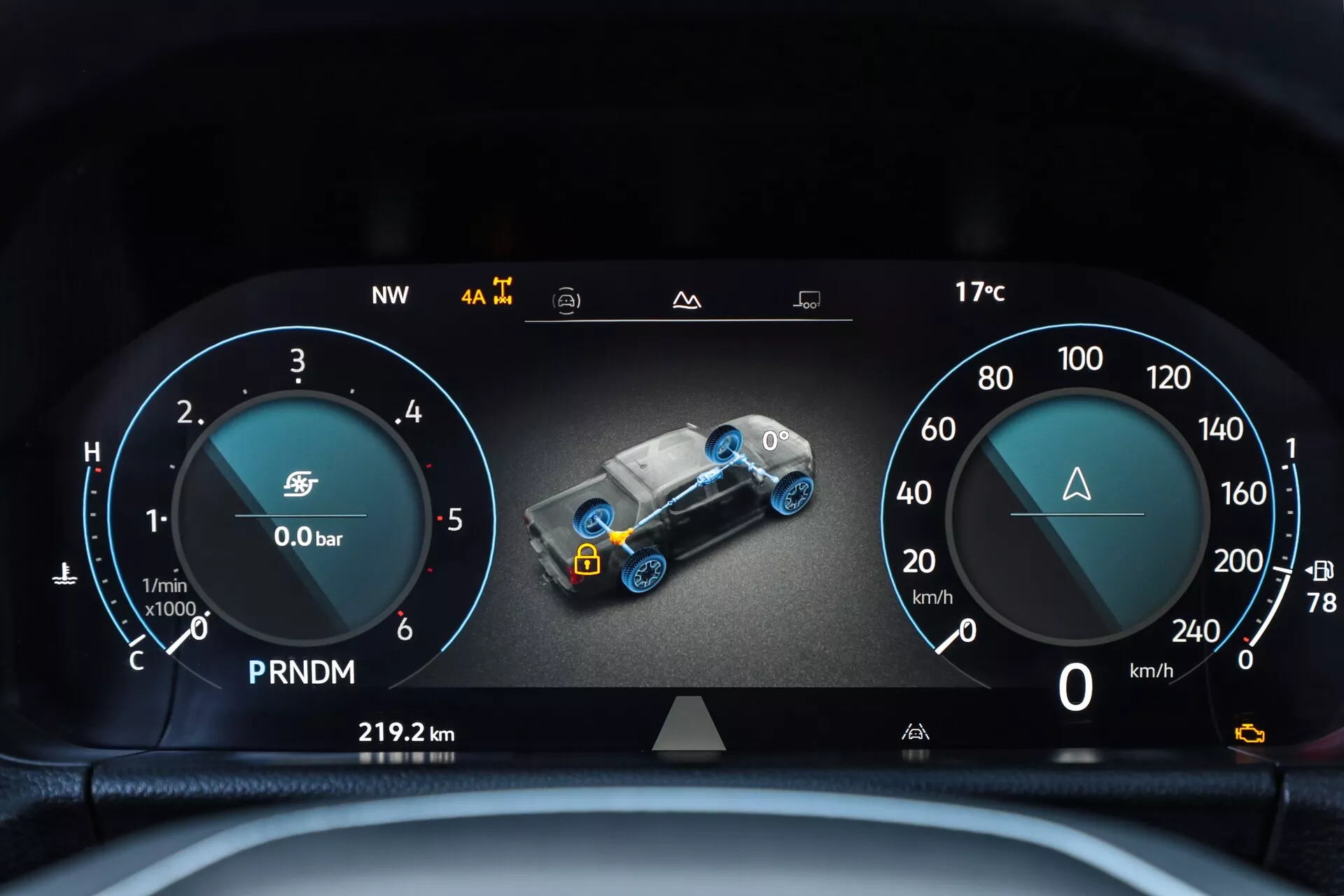 Podroben pogled na digitalni instrumentni grozd druge generacije VW Nutzfahrzeuge Amarok 2023 iz voznikove perspektive. Širokokoten digitalni zaslon ima eleganten dizajn z obratomerom in merilnikom goriva na levi, središčnim 3D grafičnim prikazom avtomobila za spremljanje sistema z indikatorji, kot so turbo tlak in stanje pogonskega sklopa, ter merilnik hitrosti na desni. Dodatni elementi vključujejo opozorilni trikotnik, indikator izbire prestav, števec prevoženih kilometrov, smer kompasa in zunanjo temperaturo. Ostra in dobro osvetljena slika se osredotoča na prikaz naprednih tehnoloških funkcij vozila v notranjosti kabine, poudarjena z zgornjim robom volana.