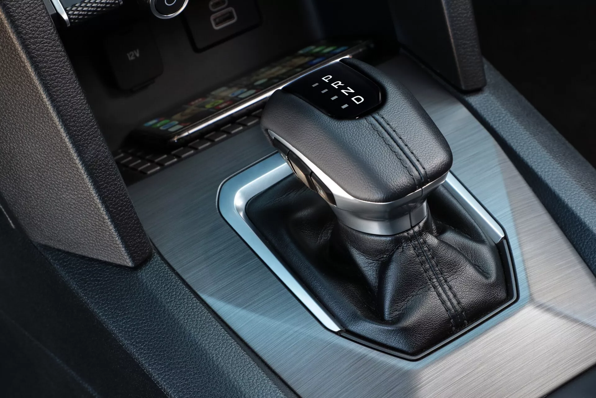 Pogled od blizu na samodejni menjalnik VW Nutzfahrzeuge Amarok 2023, s čez usnje ovitim vzvodom in osvetljenim kazalnikom prestavnega mesta, ki prikazuje 'P' za parkiranje. Kot od zgoraj in iz desnega sprednjega kota razkrije kovinski okvir z modro ambientalno osvetlitvijo in majhen predal v sredinski konzoli.