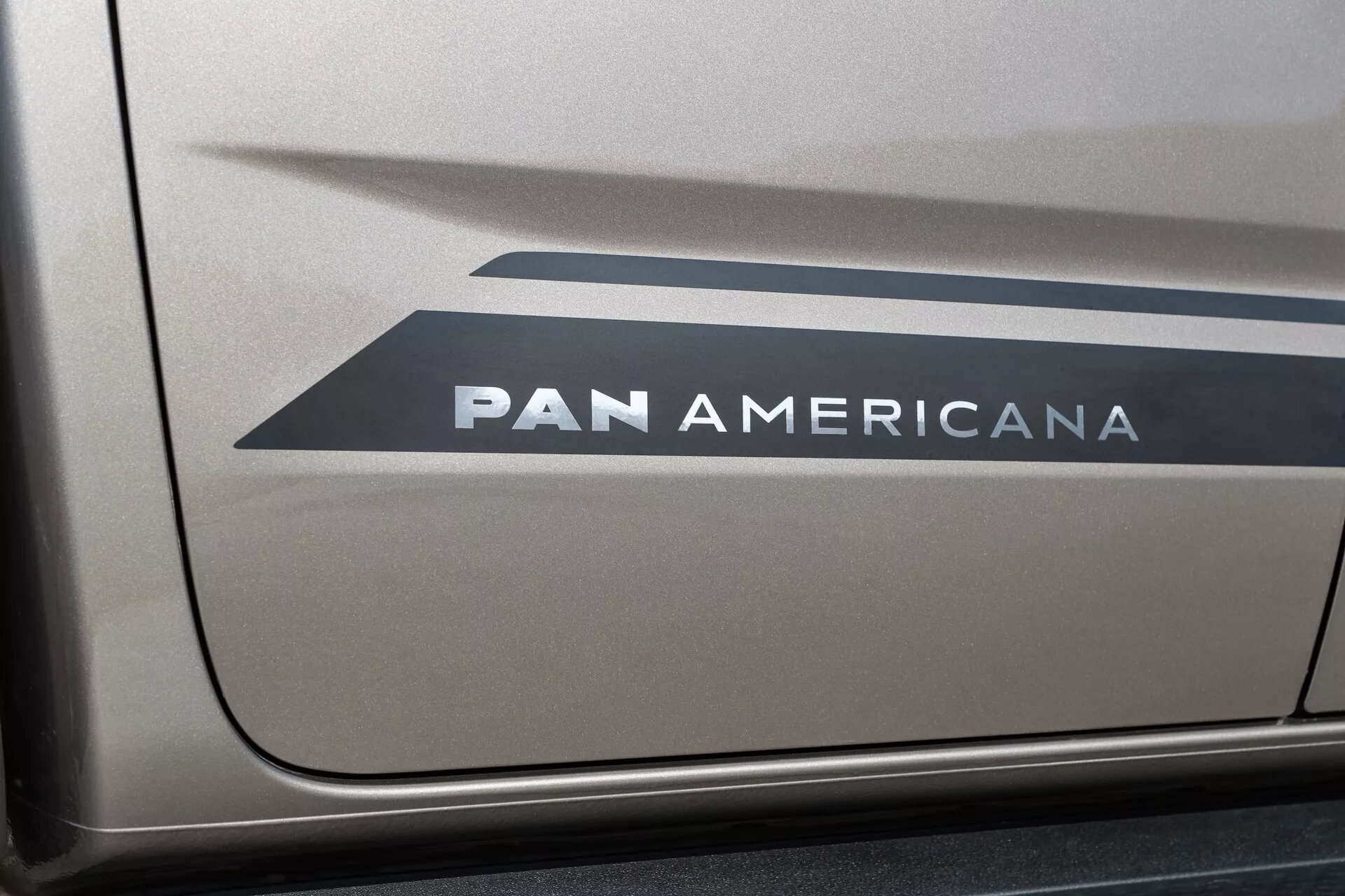 Povečan stranski pogled na del vozila VW Nutzfahrzeuge Amarok 2023 (druga generacija), ki prikazuje grafiko z dvema nazaj obrnjena črkovna znaka v obliki Chevron in odebeljeno besedilo 'PAN AMERICANA' na verjetno kovinsko barvani površini, z jasnim in enakomernim osvetlitvijo.