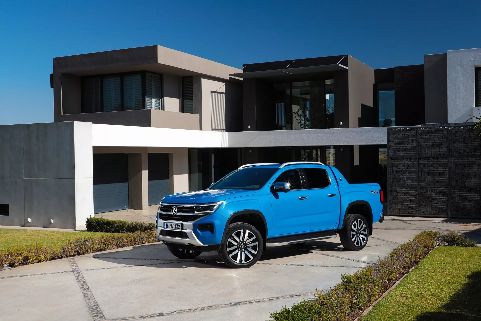 Svetlo modri drugi generacija 2023 VW Nutzfahrzeuge Amarok parkiran na betonskem dovozu pred sodobno hišo, prikazan iz trikotnega sprednjega kota. Ta pogled poudarja vidno kromirano mrežo, elegantne LED-žaromete, večkraka lita platišča, svetlečo barvo, karoserijo z dvojno kabino, ogledala z vgrajenimi smerniki in rahlo zatemnjena stekla. Prav tako sta jasno vidna visoka oddaljenost od tal in primestno okolje.