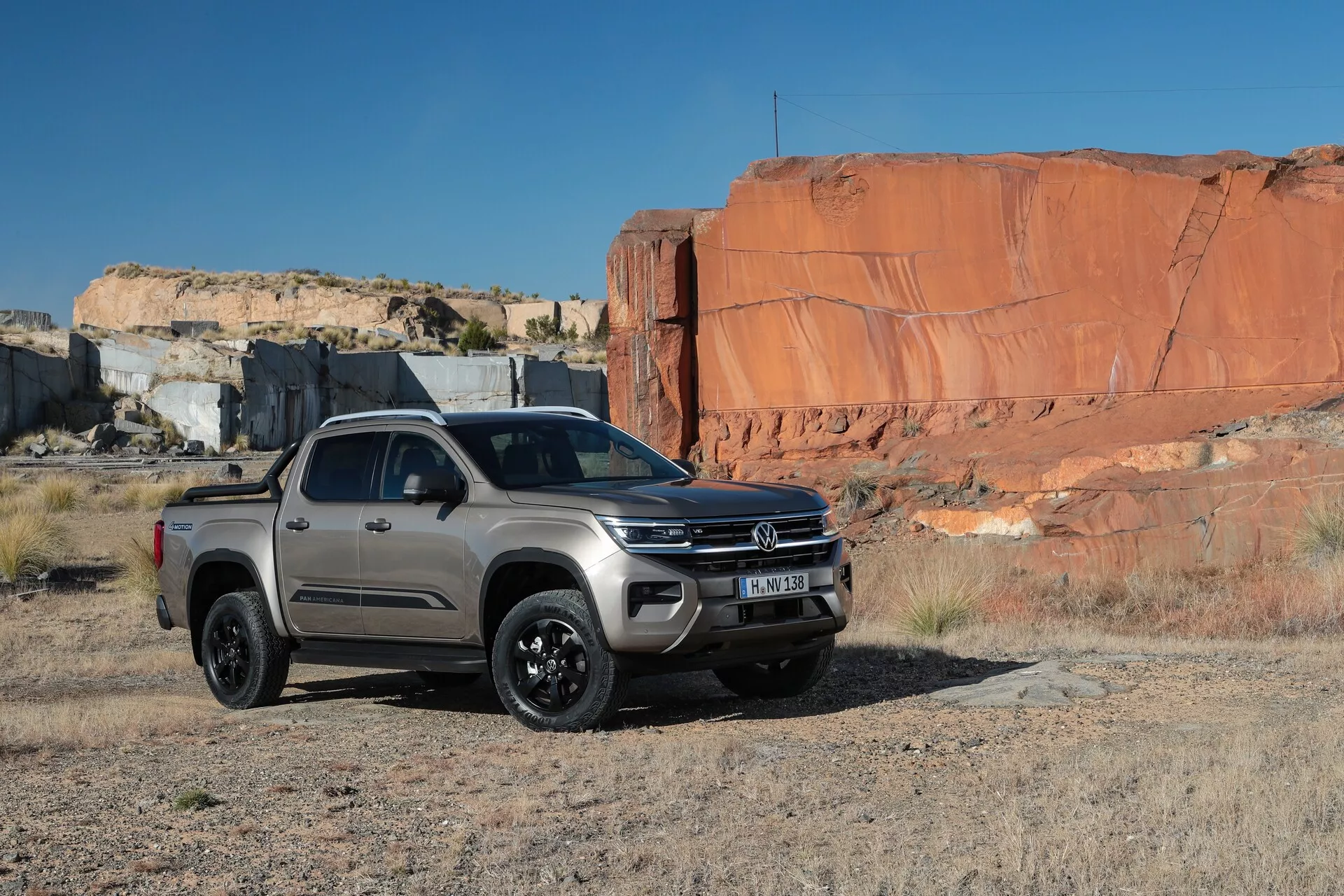 VW-Nutzfahrzeuge Amarok druge generacije iz leta 2023 prikazan na naravnem, skalnatem terenu, ki poudarja njegove terenske zmogljivosti. Posnetek iz treh četrtin spredaj prikazuje barvno usklajen sprednji odbijač z vgrajenimi meglenkami, izrazito sredinsko značko na rešetki, elegantne pravokotne žaromete, dvignjene odseke pokrova motorja, črna platišča iz lahke zlitine in visoko oddaljenost od tal. Konfiguracija z dvojno kabino ima štiri vrata z vidnimi stranskimi ogledali, kljukami vrat in stranskimi stopnicami. Tovornjak, postavljen na ravno umazano površino z ozadjem velikih rdečih skalnih formacij, je glavni objekt fotografije, ki je bila posneta pod naravno dnevno svetlobo.