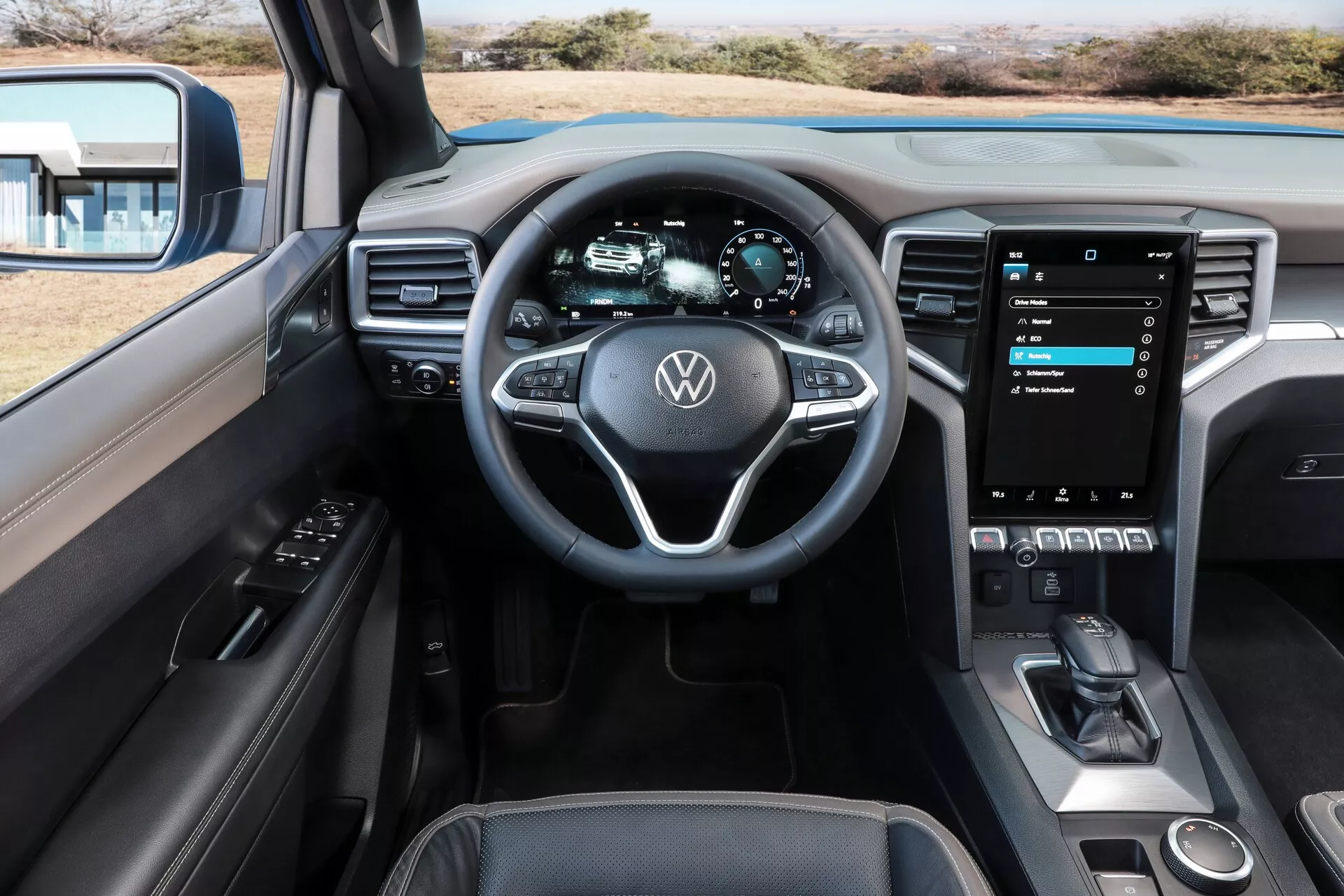 Notranji pogled s sedeža voznika v Volkswagen Amarok 2023 (2. generacija), z poudarkom na volanu z Volkswagenovim emblemom in večfunkcijskimi krmilnimi elementi, povsem digitalni instrumentni plošči, ki prikazuje meritve vozila, navpično orientiranem zaslonu na dotik infotainment sistema, občutljivih na dotik HVAC kontrolah, ročici menjalnika in razkošni estetiki kabine, ki združuje umetno usnje in kovinske poudarke.