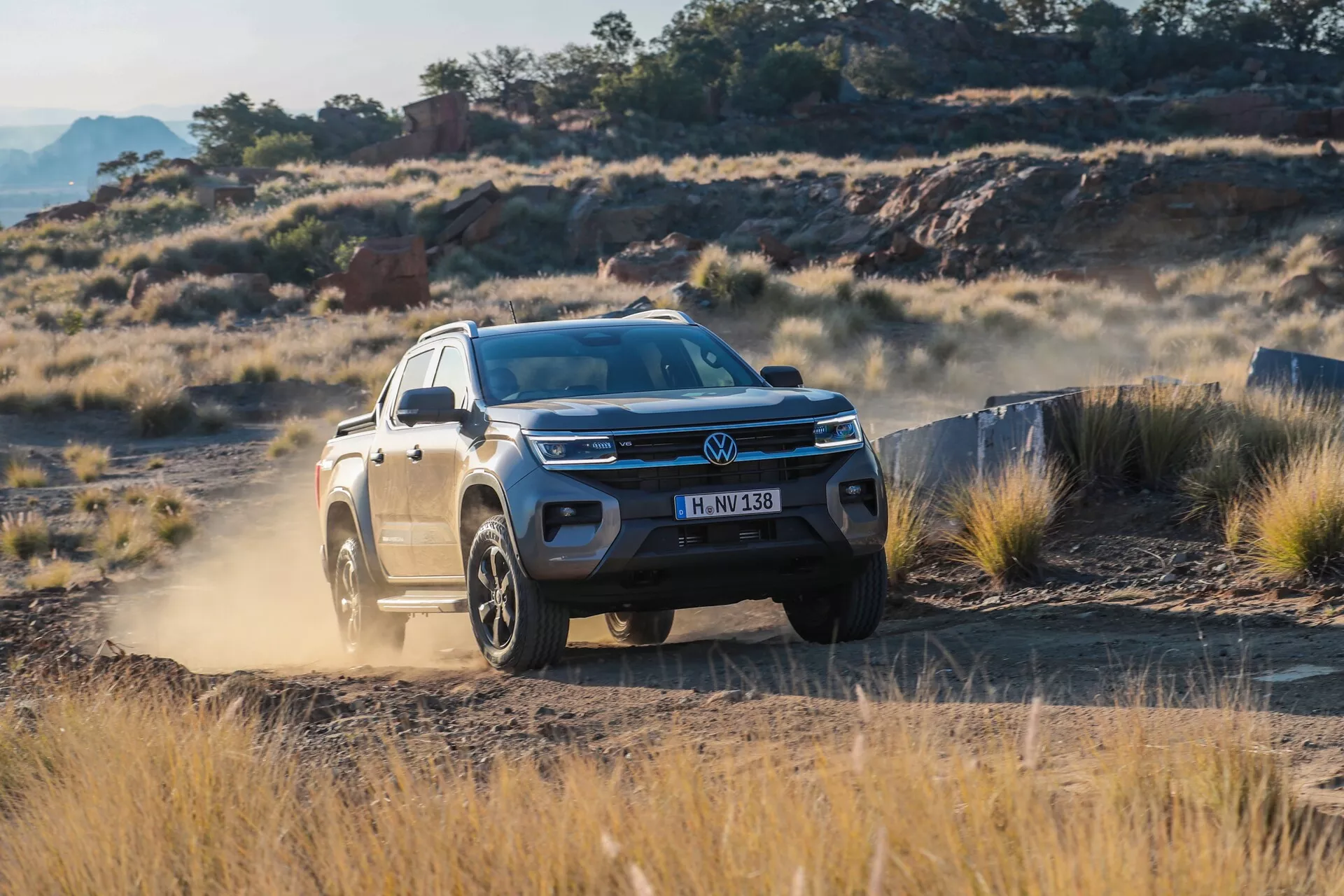 Tričetrtinski sprednji pogled na Volkswagen Amarok 2023 (2. generacija). Slika poudarja sprednjo masko z znakom Volkswagen, elegantne LED žaromete, sprednji odbijač z zračnimi režami in delno bočni profil voznikove strani, vključno s sprednjimi vrati, blatnikom, obroči koles in stranskimi ogledali. Moderni SUV z vodoravno rešetkasto masko, LED svetlobno letvijo in zlitimi kolesi za vse terene, je zajet v gibanju po skalnatem, travnatem terenu, kar prikazuje njegove terenske zmogljivosti in napredne funkcije pomoči vozniku, kot sta prilagodljiv tempomat in avtonomno zaviranje v sili.