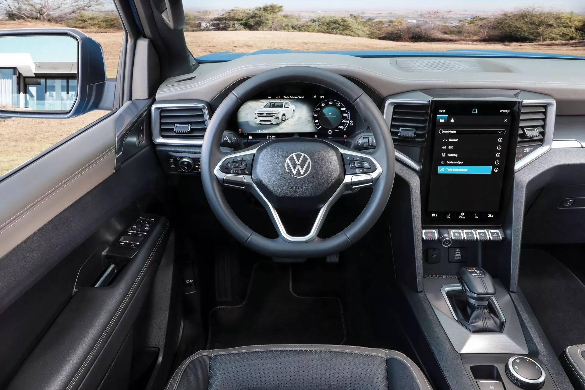 Notranji pogled na Volkswagen Amarok 2023 (2. generacija), osredotočen na voznikovo kabino iz osrednje perspektive, ki spominja na voznikov pogled. Slika prikazuje trikraki volan z večfunkcijskimi gumbi, popolnoma digitalno instrumentno ploščo, ki prikazuje osnovne podatke o vožnji, in velik, navpično usmerjen zaslon na dotik sistema za zabavo in informacije. Sredinska konzola vključuje ročico za samodejni menjalnik, motorni gumb za zagon/zaustavitev, držala za skodelice in shranjevalni predal. Notranjost ima dvoobarvno shemo z usnjem in mehkimi plastičnimi materiali, kromirane poudarke ter znake tehnološke integracije, kot so električna okna, funkcije povezljivosti in napredni sistemi za pomoč vozniku.