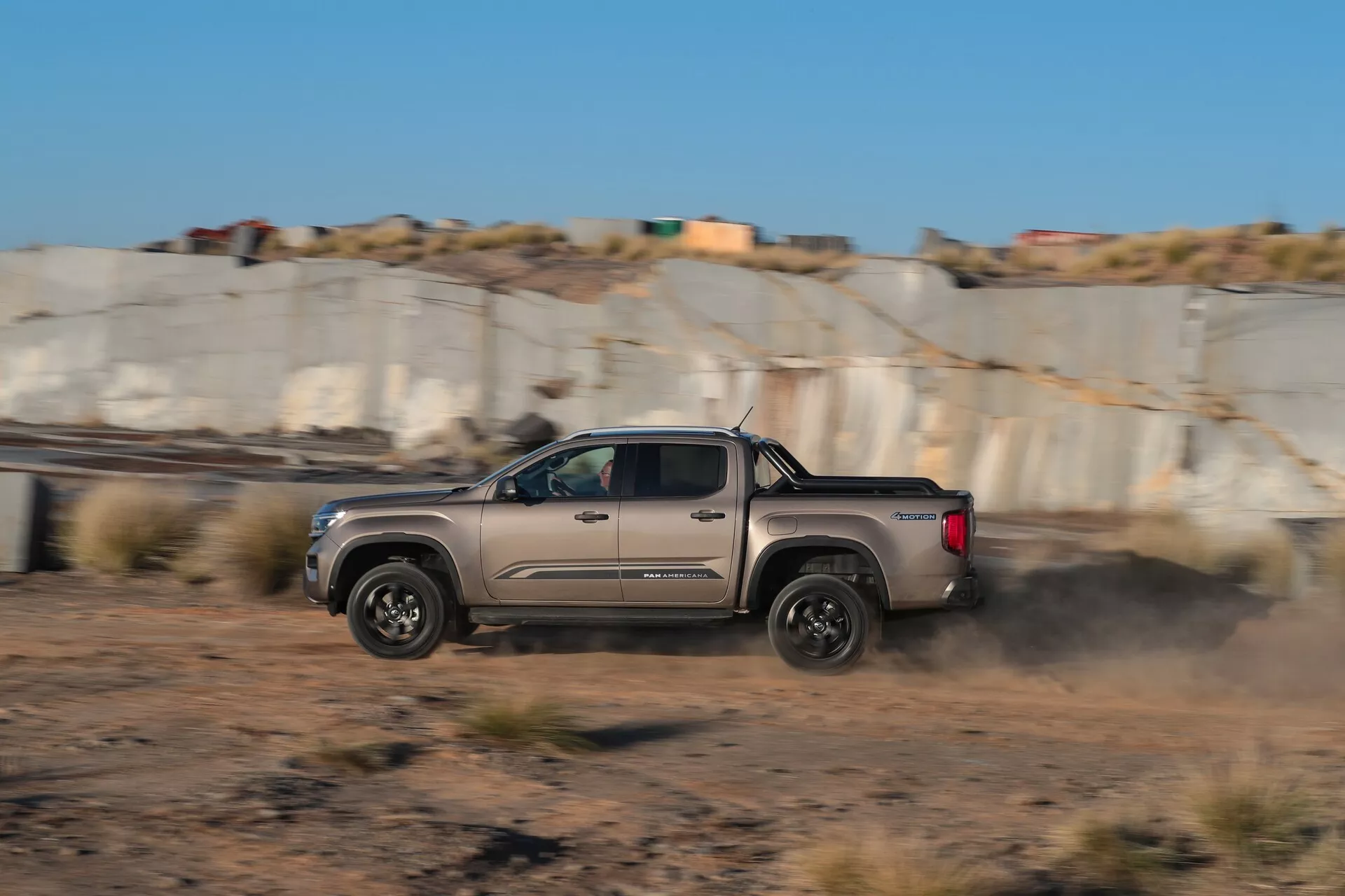 Stranski profil vozila Volkswagen VW Nutzfahrzeuge Amarok 2. generacije 2023, ujetega v gibanju z zabrisanim ozadjem in oblakom prahu za njim, kar poudarja njegov dinamični manever po makadamski cesti. Ima dvojno kabino, lita platišča in aerodinamično sodobno obliko, kar nakazuje napredne terenske zmogljivosti.