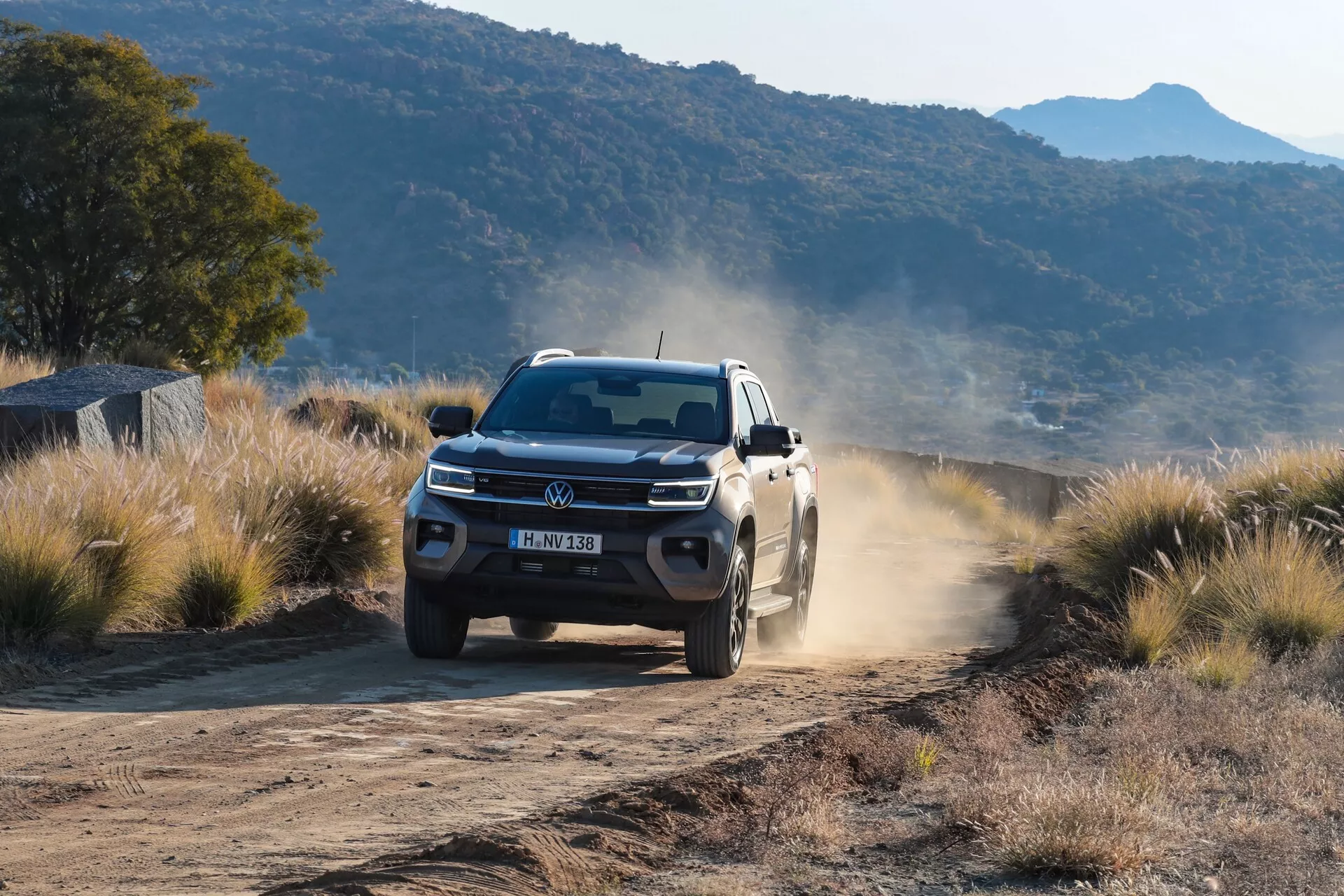 Beli pickup Volkswagen Amarok 2023 (2. generacija) ujet v gibanju na neasfaltirani, prašni podeželski cesti, ki dokazuje svoje terenske sposobnosti. Vozilo je prikazano z nizkega, sprednjega kota, poudarjajoč robustno zasnovo, LED žaromete, embleme VW, aluminijasta platišča in prah, ki ga pušča za seboj. Scena namiguje na zgodnje jutro ali pozno popoldne z daljšimi sencami pod vozilom.