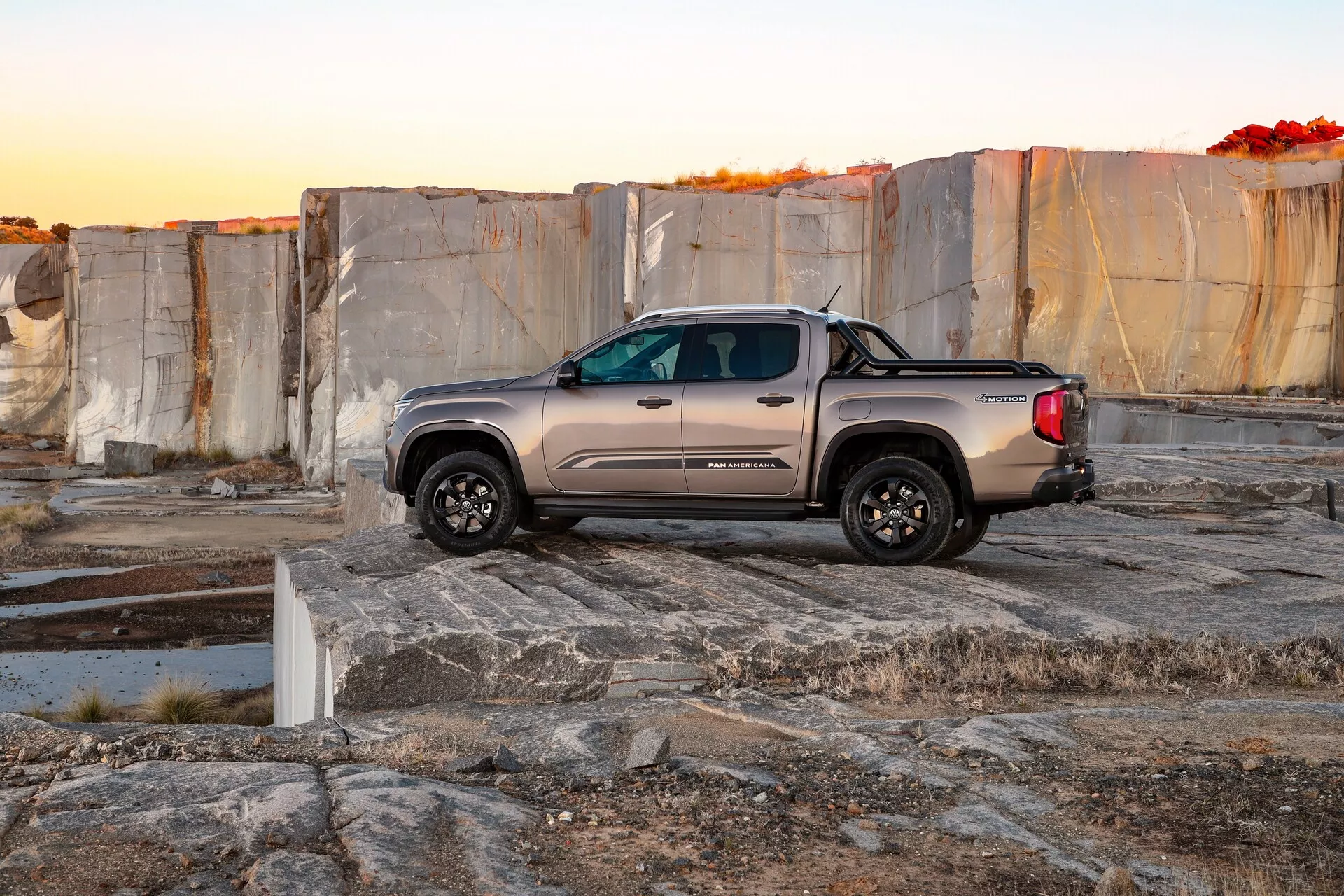 Fotografija poltovornjaka VW Nutzfahrzeuge Amarok iz leta 2023 na prostem, verjetno v kamnolomu ali območju z velikimi kamnitimi ploščami. Slika ujame tovornjak ob zori ali mraku, poudarjajoč njegovo aerodinamično karoserijo, dvojno kabino, robustna terenska kolesa, kromirane poudarke in kovinsko barvo. Tovornjak je postavljen diagonalno, s sprednjim delom rahlo nagnjenim proti kameri, prikazujejoč njegova zunanja vzvratna ogledala, nastavljive značilnosti in lita kolesa. Puščavsko okolje in neenakomerna kamnita površina poudarjata trpežnost in terenske zmogljivosti vozila.
