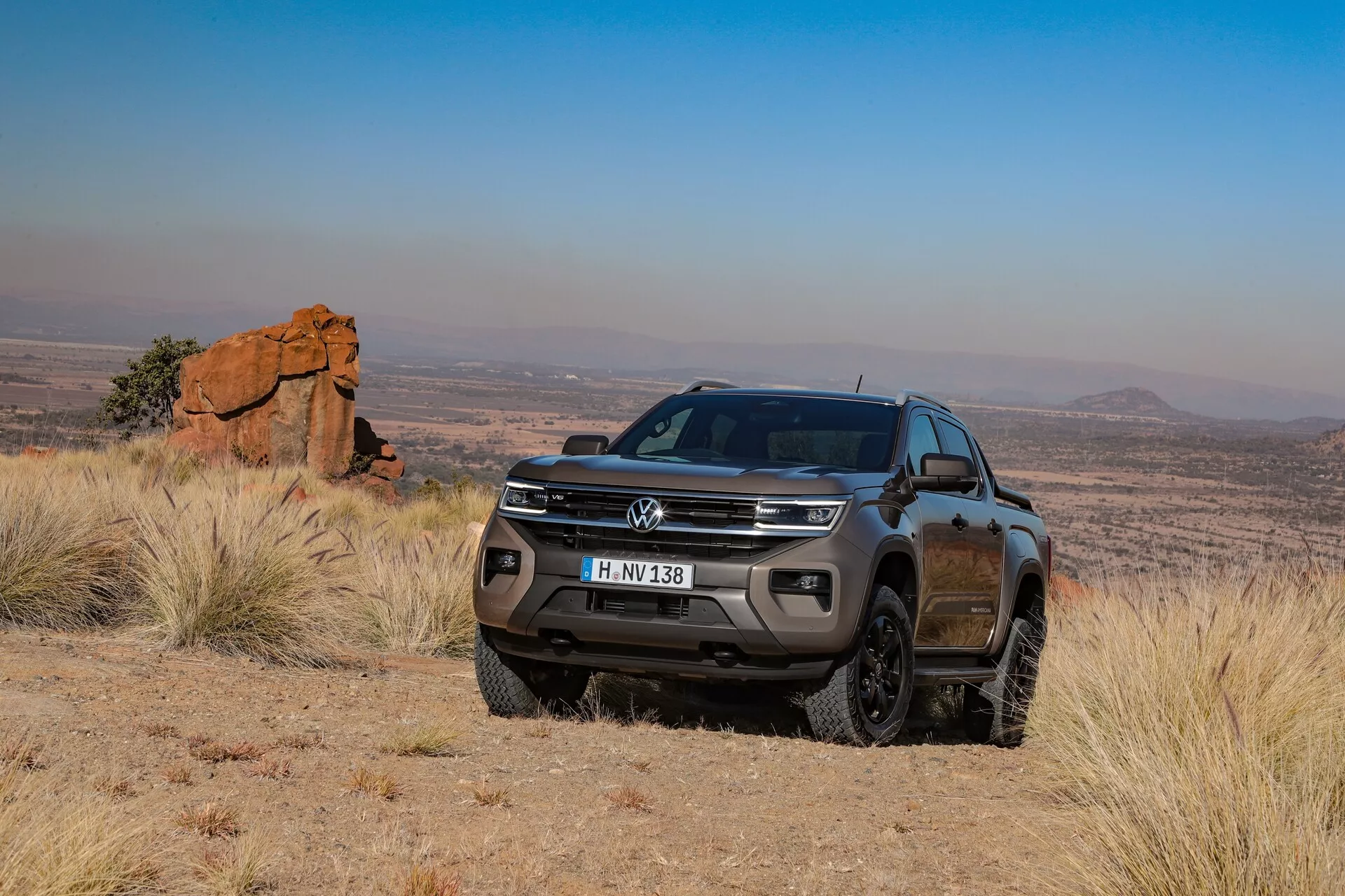 Volkswagen Amarok 2023 (2. generacija) poltovornjak v zunanjem, terenskem okolju, ujeta v tričetrtinskem sprednjem kotu, prikazuje svojo potniško stran s jasnimi pogledi na sprednjo mrežo, odbijač, žaromete, meglenke, oboke koles in del stranskega profila, vključno z vrati za potnike in stranskim ogledalom. Poudarjena drža, velika oddaljenost od tal in robusten tek pnevmatik poudarjajo njegove terenske zmogljivosti. Slikovito ozadje grobega terena in oddaljenih hribov še poudari obliko in pustolovsko naravo vozila.