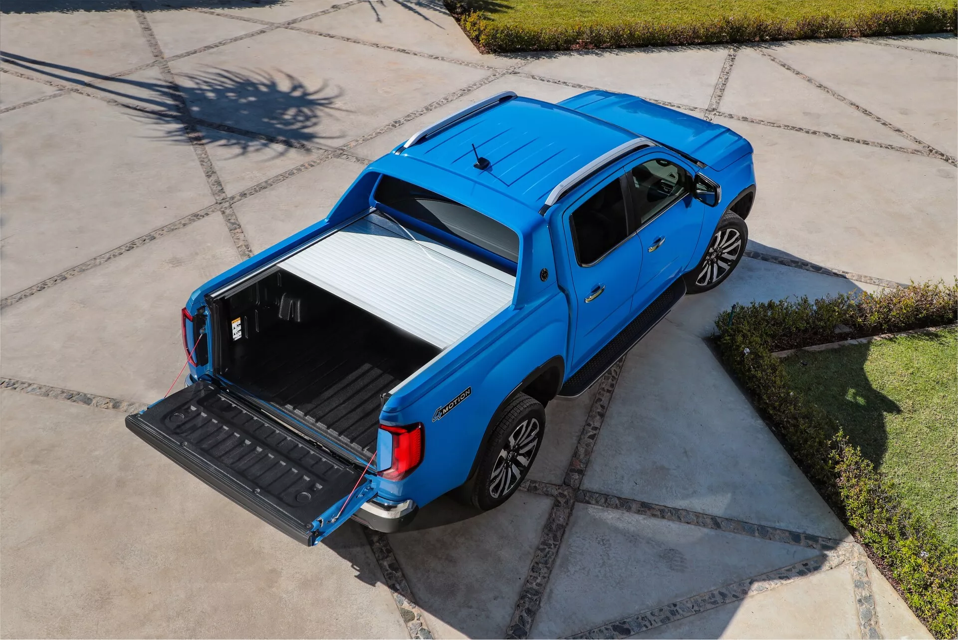 Zračni posnetek modrega pickup VW-Nutzfahrzeuge Amarok 2. generacije za leto 2023, ki prikazuje njegov zgornji in bočni profil iz visoke perspektive. Vozilo je parkirano na značilno tlakovani dovozni poti, obsijano s sončno svetlobo. Posnetek iz rahlo zadnje poševne perspektive poudarja odprt tovorni prostor z znižanim zadnjim vrati, ki razkriva vzdržljivo, zaščitno oblogo. Eleganten, sodoben štirivratni dizajn kabine poudarjajo stranska ogledala in kljuke vrat v barvi karoserije. Kolesa imajo večkraki oblikovanje iz lahke zlitine, ki izboljšuje tako estetiko kot potencialno zmogljivost.