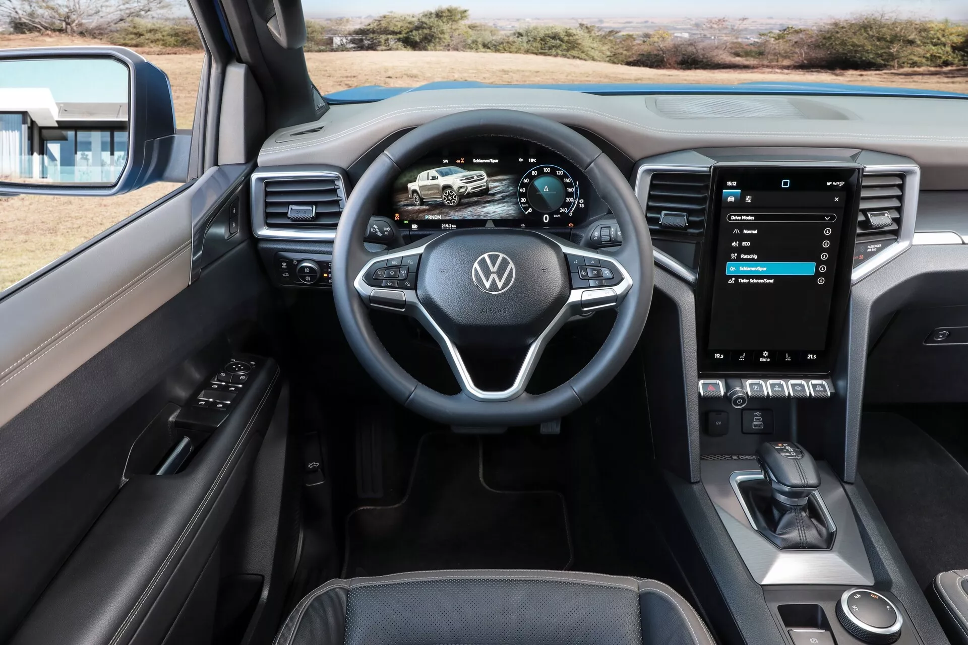 Slika prikazuje notranjost vozila Volkswagen Nutzfahrzeuge Amarok 2023 (2. generacija) s perspektive prednjega potnika. Osredotoča se na večfunkcijski volan z logotipom Volkswagen. Za njim je popolnoma digitalna instrumentna plošča s prikazom hitrosti, vrtljajev in navigacije. Osrednjo konzolo dominira velik, pokončno nameščen zaslon na dotik za informacije in zabavo, ki ponuja nastavitve vozila, avdio nadzor in povezljivost pametnega telefona, kot sta Apple CarPlay ali Android Auto. Spodaj so vidni minimalni fizični gumbi, elegantna prestavna ročica, odprt prostor za shranjevanje in brezžična polnilna plošča, kar nakazuje na sodoben, napreden dizajn. Notranjost izžareva čisto in sodobno estetiko z mehkimi površinami na dotik, brušenimi kovinskimi poudarki in centralnim zvočnikom, kar kaže na premium avdio sistem.