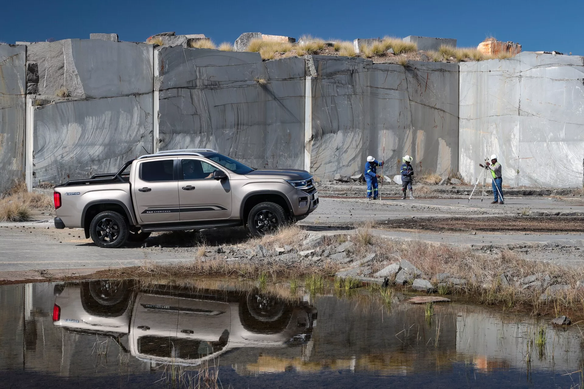 Srednje velik pickup Volkswagen Nutzfahrzeuge Amarok 2023 v konfiguraciji crew cab je parkiran pod poševnim kotom z sprednjim delom, obrnjenim na desno. Stranski profil razkrije štiri vrata, stranska ogledala, kljuke na vratih, kolesa in oblikovne linije na karoseriji v nevtralni srebrni ali svetlo sivi barvi s kontrastnimi črnimi poudarki okoli kolesnih lokov in spodnjega dela karoserije. Vozilo stoji na gramozni ali zemeljski površini s krpami trave in lužo, ki odseva njegov spodnji del. V ozadju je kamnolom ali rudniška lokacija z velikimi kamnitimi ploščami ter delavci s čeladami in odsevnimi telovniki, kar kaže na industrijsko ali gradbeno okolje. Pomembni tehnološki elementi vključujejo aluminijasta kolesa in zunanja ogledala z veliko oddaljenostjo od tal. Slika je posneta podnevi z jasnim naravnim osvetlitvijo.