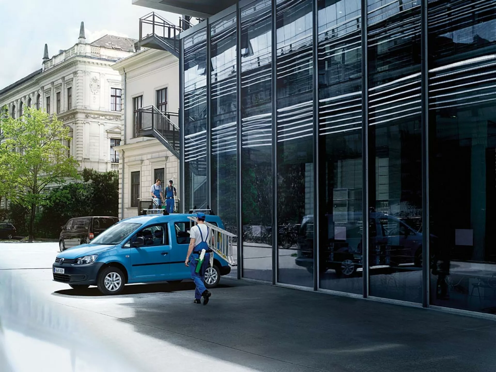Modri VW Caddy letnik 2010 parkiran na mestni ulici, prikazan iz treh četrtinskih sprednjih pogledov z odprtimi vrati in poudarjanjem njegove sprednje rešetke, žarometov, koles in vpogleda v notranjost proti ozadju moderne in klasične arhitekture