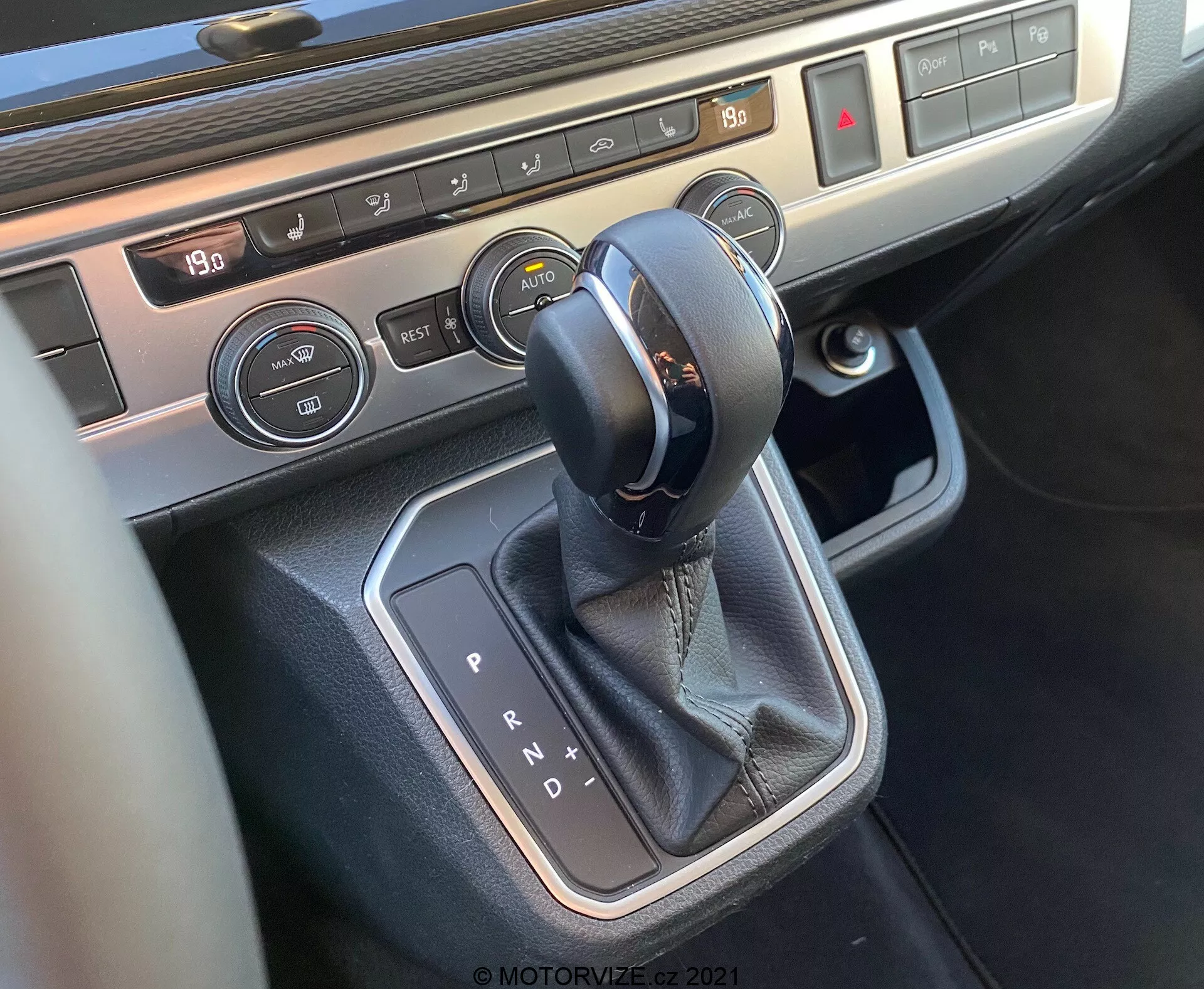 Podroben pogled na središno konzolo vozila VW Nutzfahrzeuge T6-1 Multivan 2019, osredotočen na območje prestavne ročice. Sliko prikazuje avtomatski selektor prenosa z usnjenimi šivi, obdan s kovinsko obrobo, gumbe za nadzor klimatske naprave s prikazom temperature in nadzorom za ogrevanje sedežev, kar poudarja moderno notranjo tehnologijo in vrhunske materiale vozila.