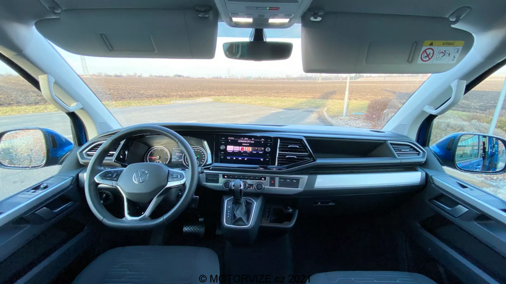 Notranjost VW Nutzfahrzeuge Transporter 2019 (T6.1 Multivan), posneta iz zadnjega sedeža proti sprednji armaturni plošči. Fotografija je posneta podnevi z naravno svetlobo, ki vstopa skozi sprednje vetrobransko steklo, in ponuja podroben pogled na prostor kabine, prednje sedeže, volan z vgrajenimi kontrolami, osrednji zaslon na dotik za zabavno-informacijski sistem, gumbe in gumbe klimatske naprave, avtomatsko prestavno ročico, shranjevalne prostore, priključke za povezavo, upravljalnike za vrata in ogledala, varnostne funkcije in udobno zasnovo sedežev.