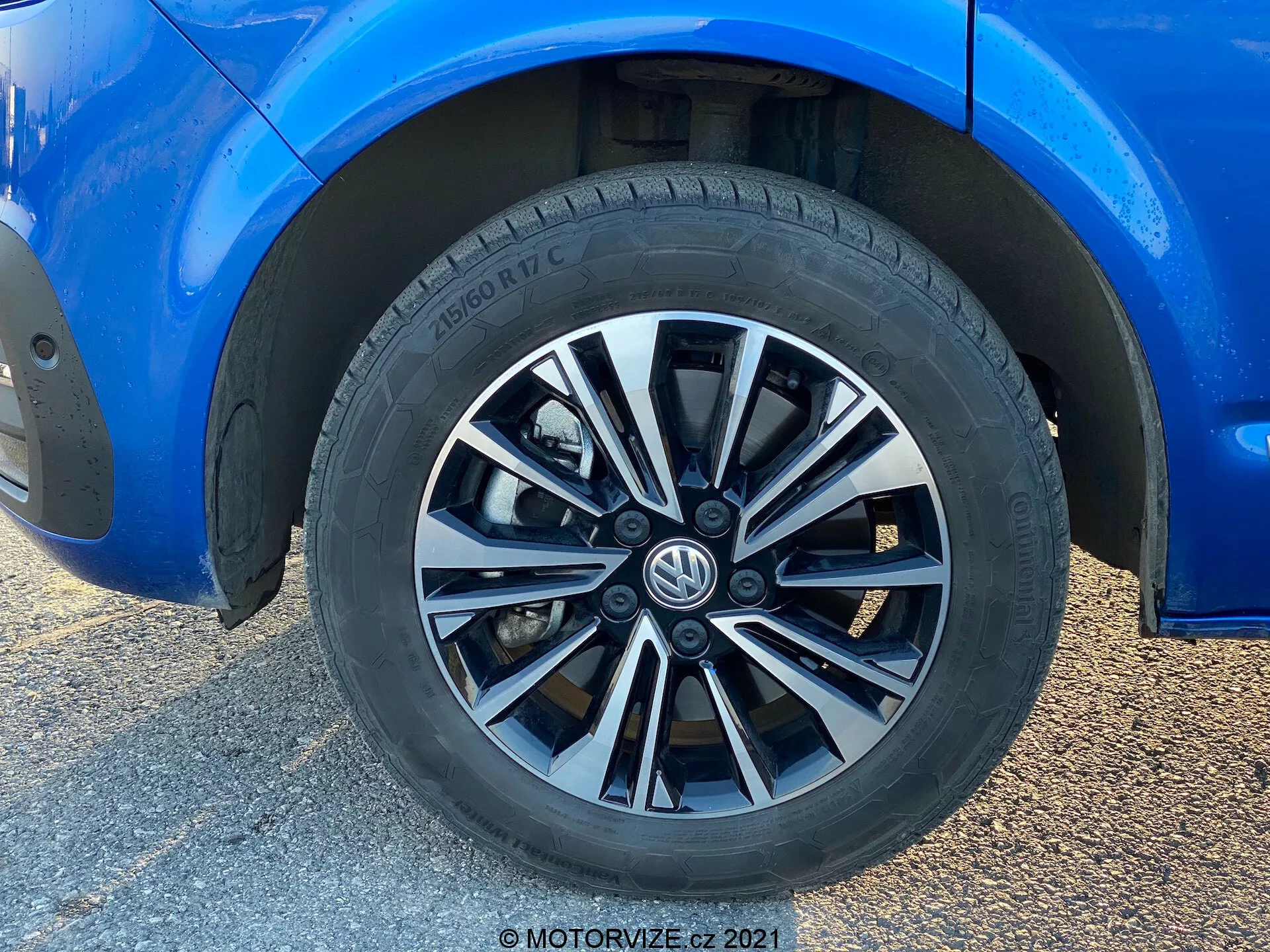 Pogled od blizu na kolo Volkswagen T6-1 Multivan 2019. Ta posnetek polnega kadra prikazuje večkraki dizajn kolesa z menjavanjem srebrnih in temnih zaključkov. Pnevmatika znamke Hankook prikazuje vzorec za cestno zmogljivost in delne specifikacije pnevmatike. Obdana modra karoserija je v čudovitem stanju in kaže odsevni sijaj. Je zmerna višina od tal in delno vidni standardni zavorni čeljusti in rotorji. Naravna svetloba poudari kompozicijo, osredotočeno na kolo, zamegljeno ozadje pa je posledica posnetka od blizu pod nizkim kotom.