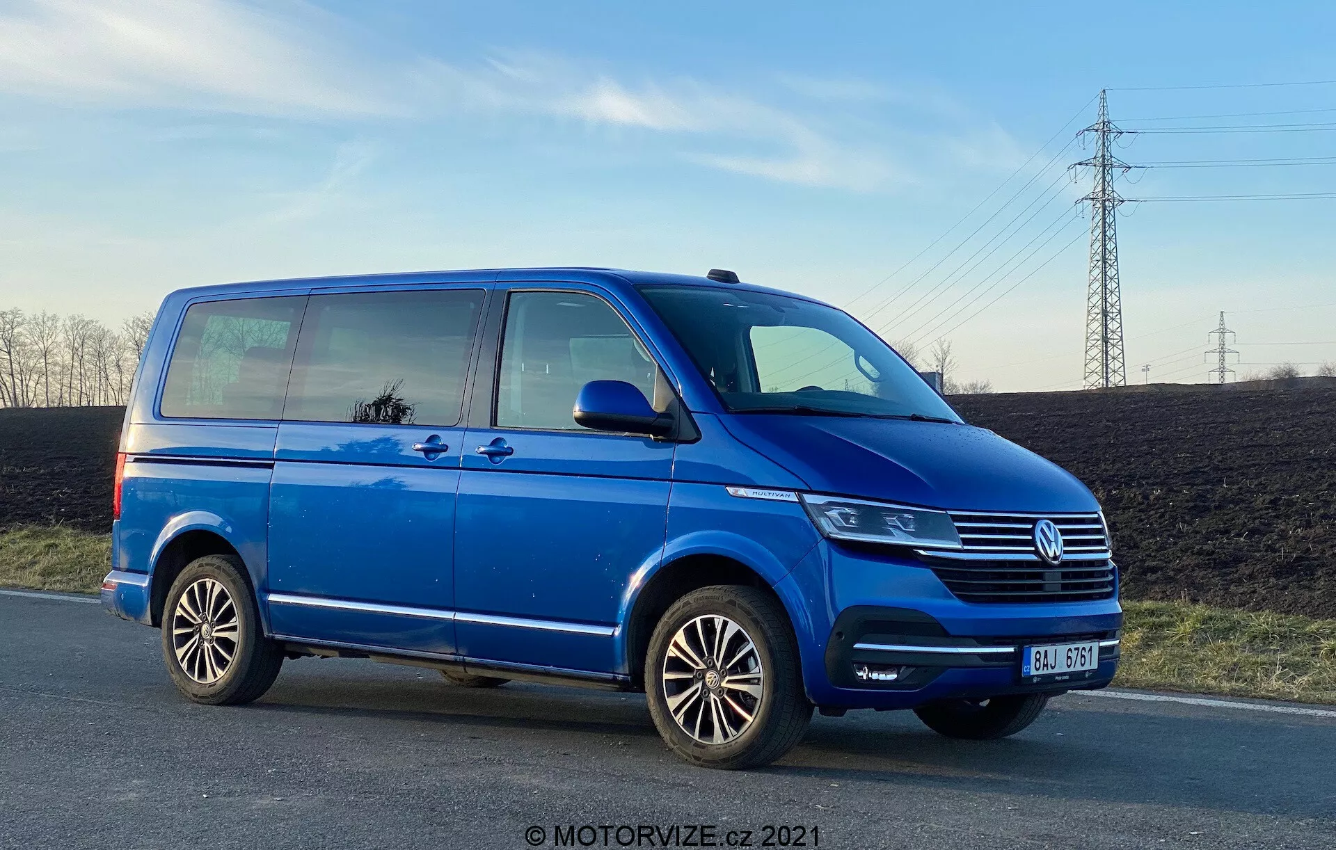 Modri Volkswagen Transporter T6 kombi, ki prikazuje njegov stranski profil z nizke kote, poudarjajoč njegovo dolžino in višino. Slika prikazuje podrobnosti, kot so drsna vrata za potnike, sprednja vrata za potnike, zadnji kolo na strani potnika, več-zračna lita platišča ter delni prikazi žarometa, Volkswagenove maske z emblemom in sprednjega odbijača. Fotografija, posneta v poznih popoldanskih urah, prikazuje oddaljenost od tal in odseva nebo in okolico v oknih, s čistim nebom in električnimi žicami v ozadju.